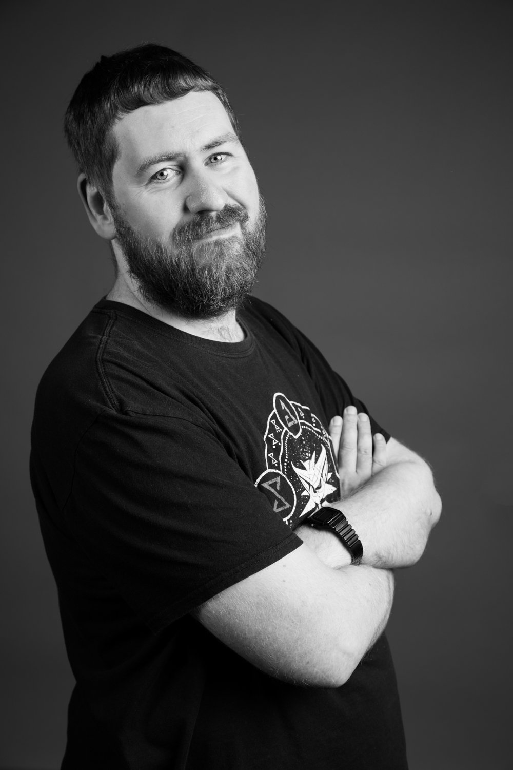 A black and white studio portrait of a man with a beard, short hair, crossing his arms, wearing a T-shirt with a graphic design and a wristwatch, smiling slightly.