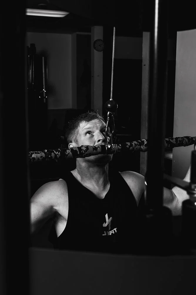 A man in workout attire performing a weightlifting exercise at the gym, with a pulley system overhead in black and white.