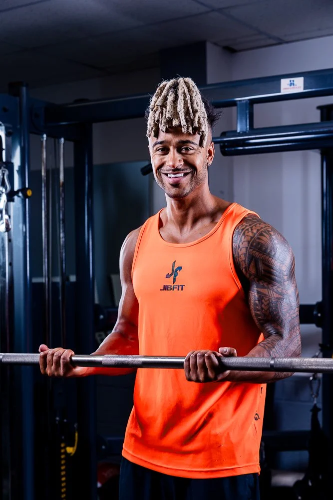A smiling man with blonde dreadlocks in an orange tank top holding a barbell in a gym.