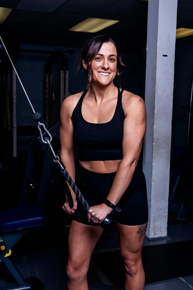 A fit woman with short brown hair smiling at the gym, wearing a black sports bra and shorts, holding a cable handle, with gym equipment and a concrete pillar in the background.