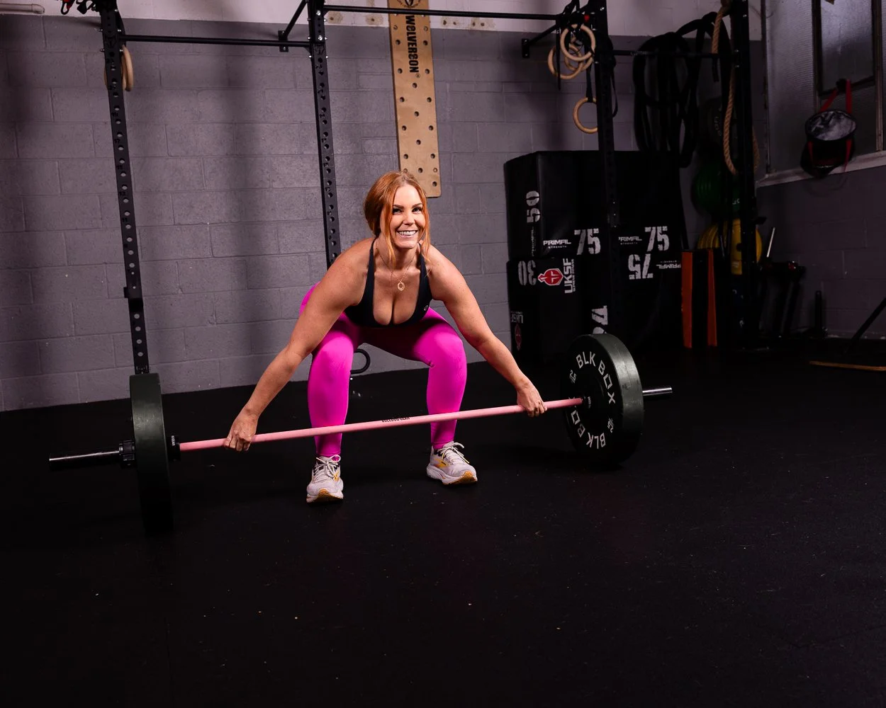 A woman lifting a barbell with weights in a gym, crouching down with a smile, wearing pink leggings and a black sports top.