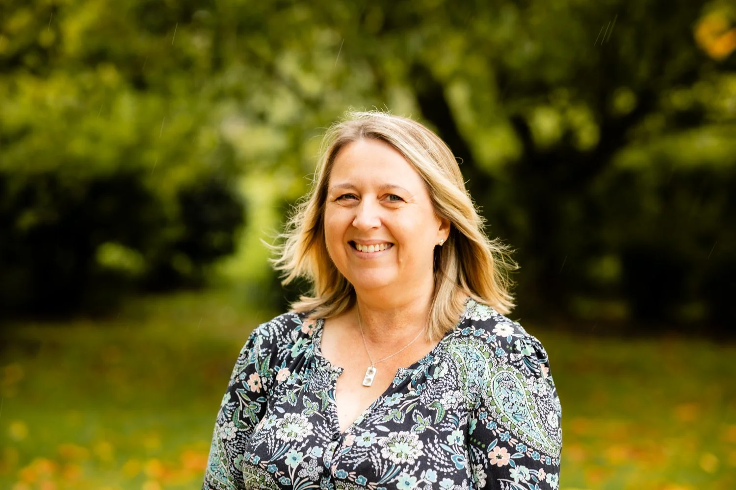 Smiling woman with shoulder-length blonde hair wearing a floral patterned blouse and a necklace, standing outdoors in a green park with blurred trees in the background.