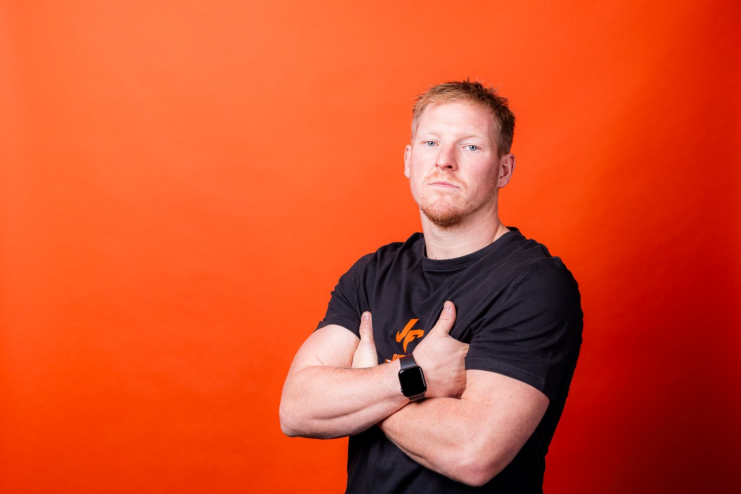 A young man with red hair and fair skin standing confidently with arms crossed, wearing a black t-shirt and smartwatch, against an orange background.