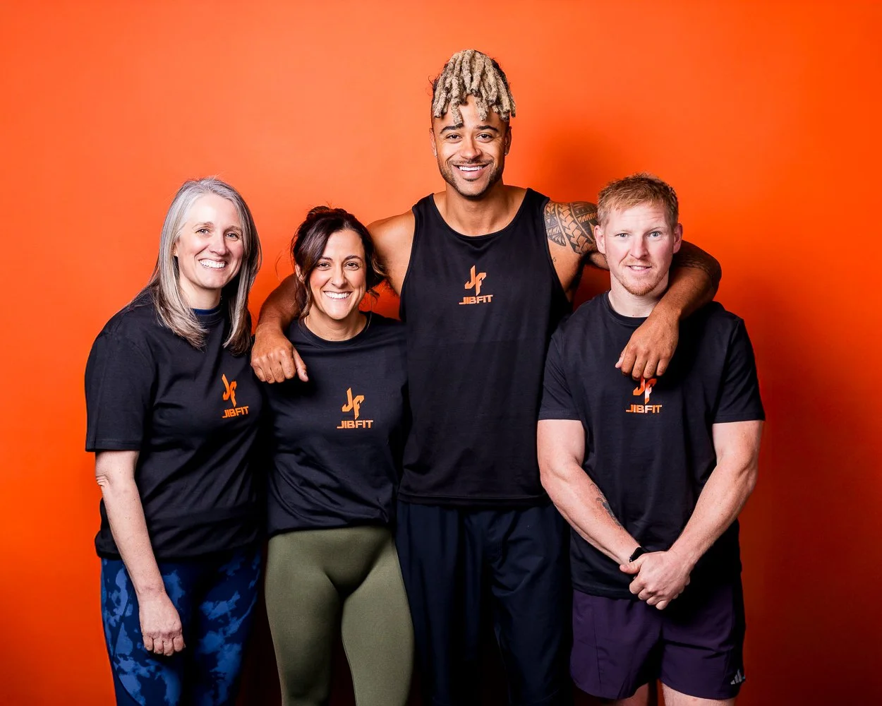 Group of four people wearing black JIBFIT shirts, standing together against an orange background. They are smiling and appear to be fitness enthusiasts or instructors.