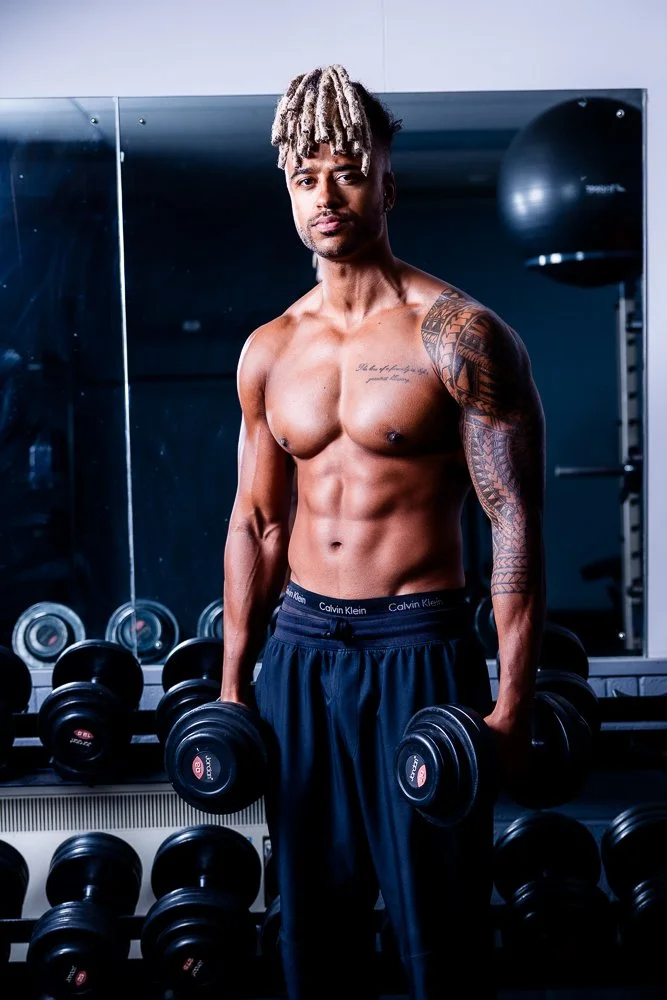 A shirtless man with tattoos on his left arm and chest, holding dumbbells in a gym with mirrors and dumbbell racks in the background.
