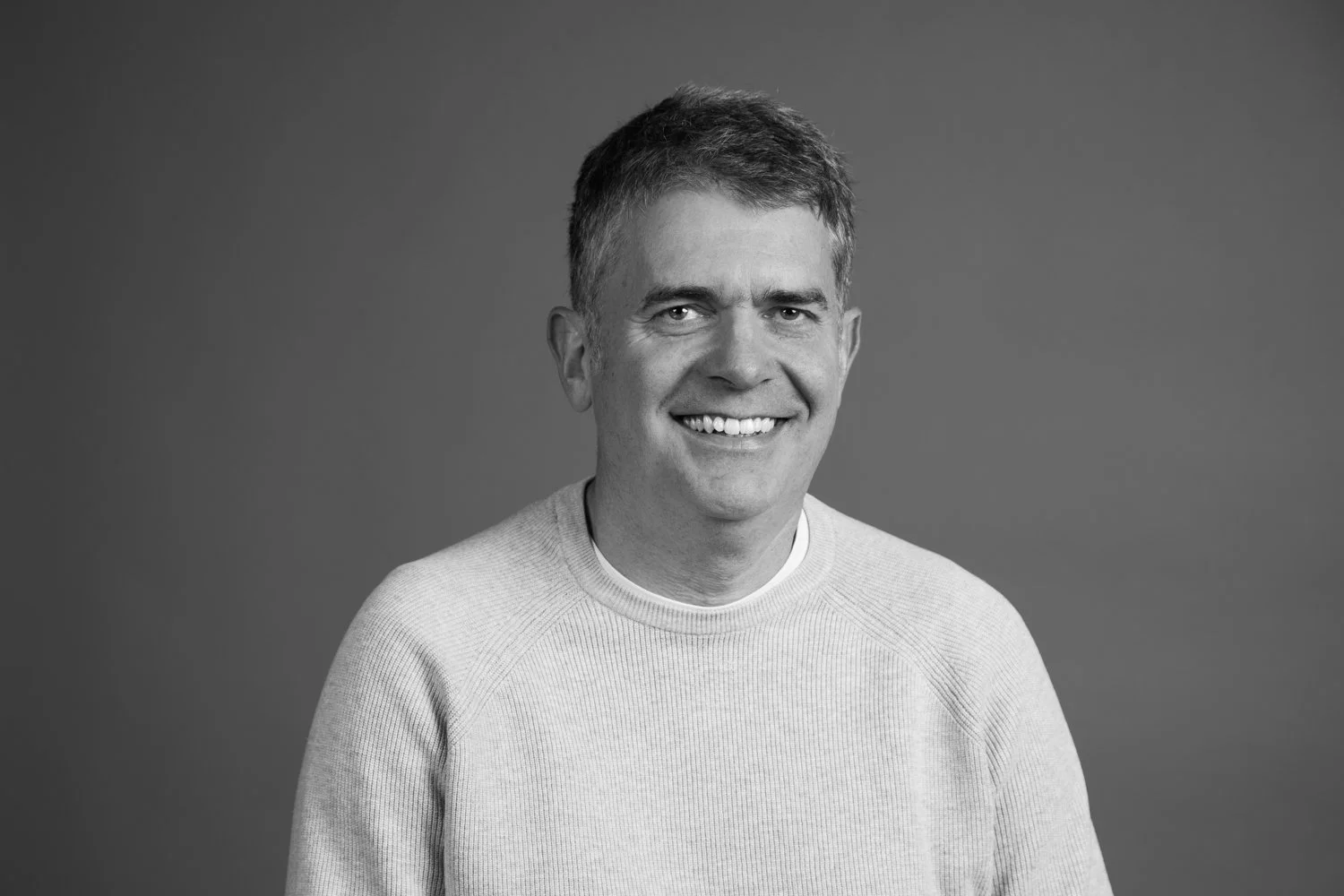 Black and white portrait of a smiling man with short hair wearing a light-colored sweater against a plain background.
