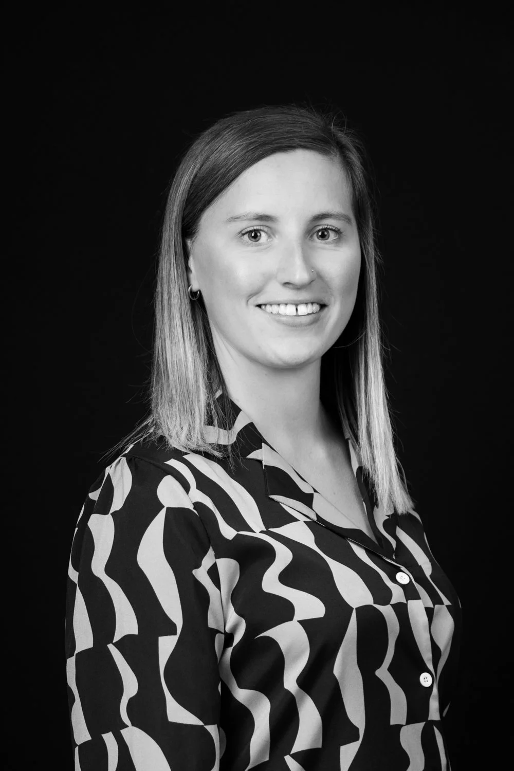 Black and white portrait of a young woman with shoulder-length hair, smiling, wearing a patterned shirt, against a dark background.