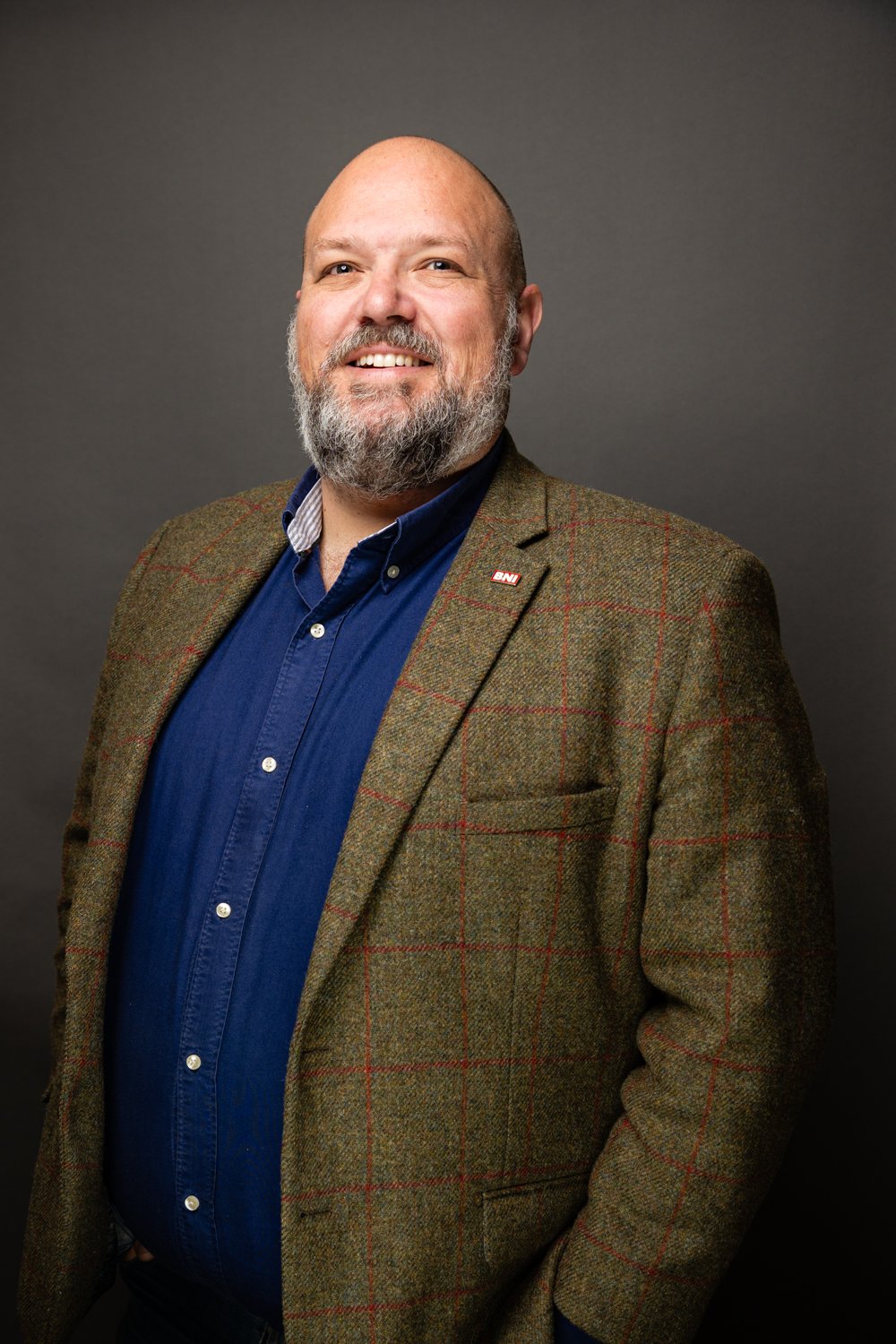 A smiling man with a beard wearing a brown plaid blazer and a blue shirt, standing against a dark gray background.