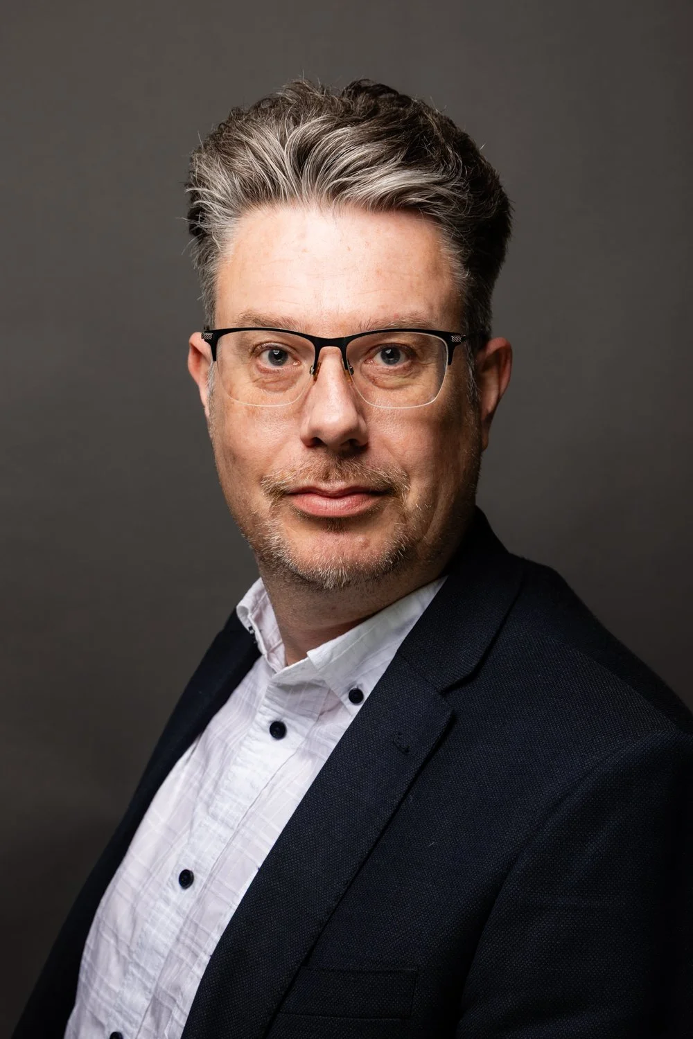 Portrait of a middle-aged man with gray hair, glasses, wearing a dark suit and white shirt, against a gray background.