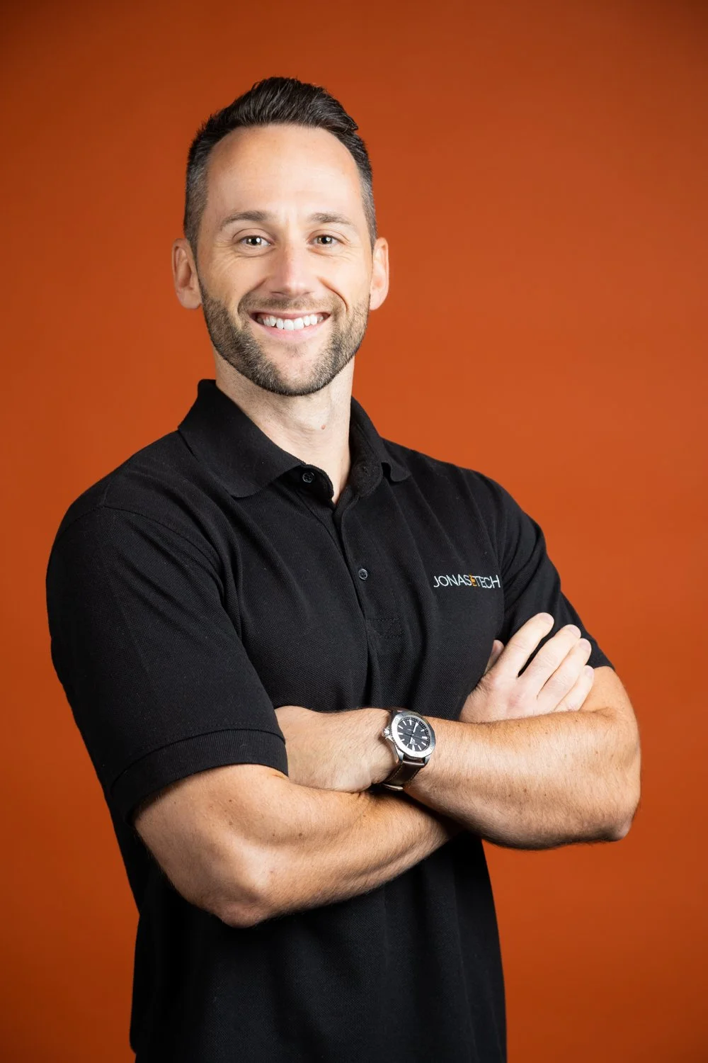 A smiling man with short dark hair and a beard, wearing a black polo shirt with a logo that reads Jonastech, standing with his arms crossed against an orange background.