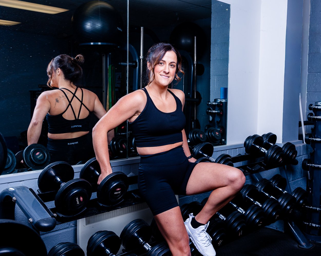 Woman in athletic wear sitting on a workout bench in a gym, holding a dumbbell, smiling, with a mirror and gym equipment in the background.