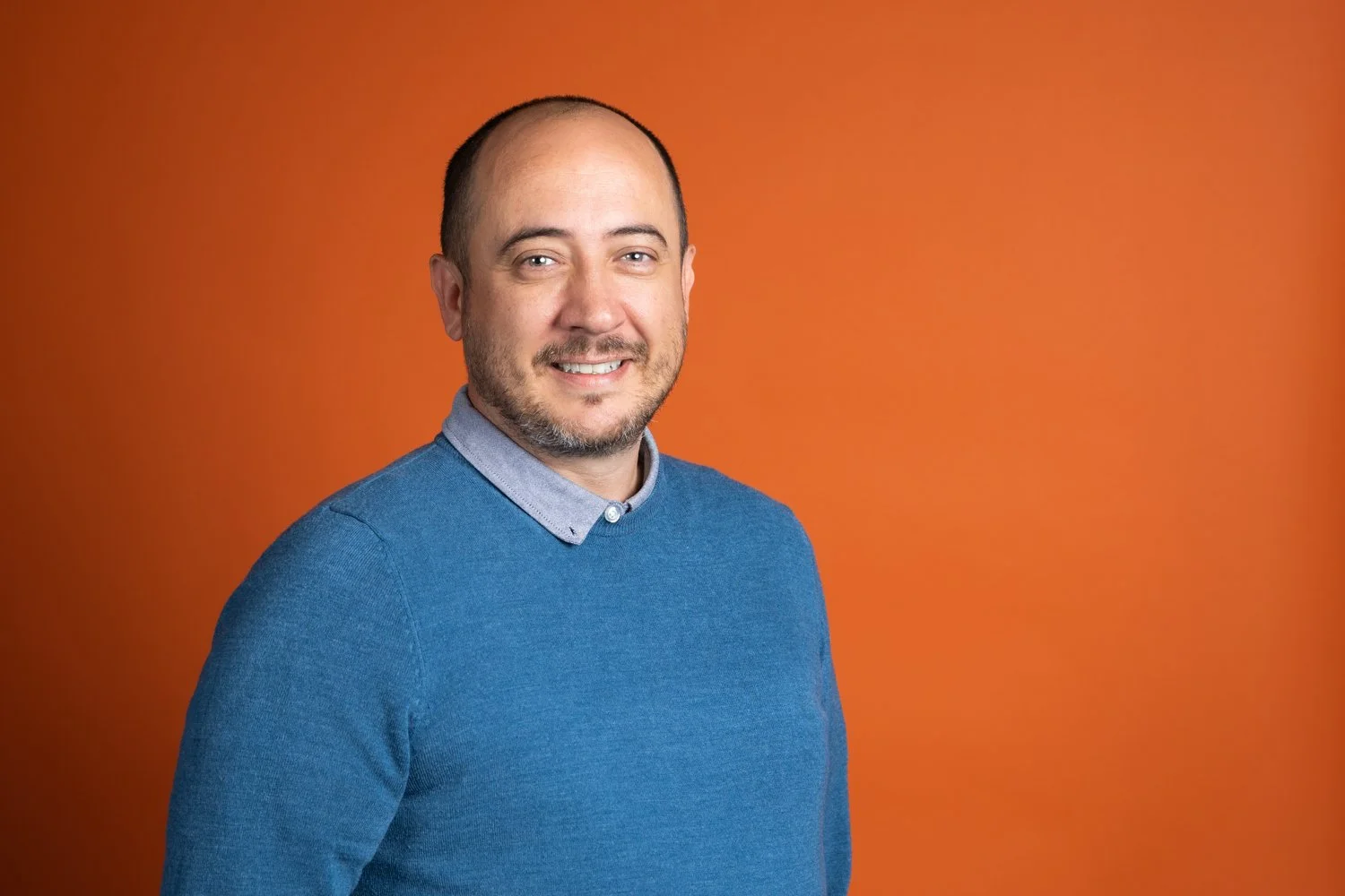 A man with a beard and short hair, wearing a blue sweater over a collared shirt, smiling in front of an orange background.