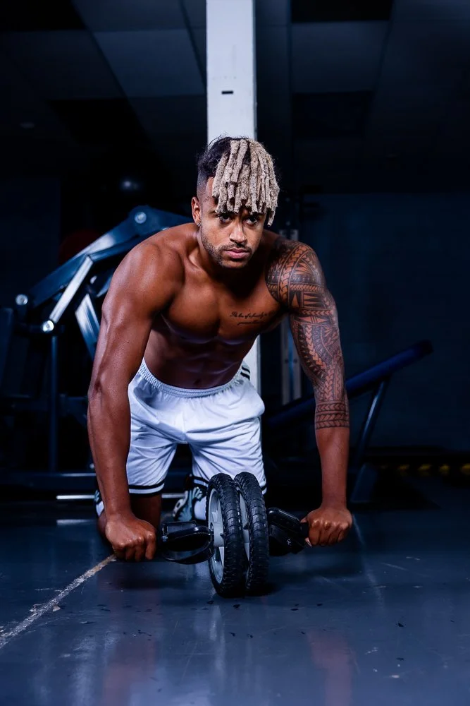A shirtless man with blond dreadlocks and tattoos on his arm is doing push-ups on an ab roller in a gym.