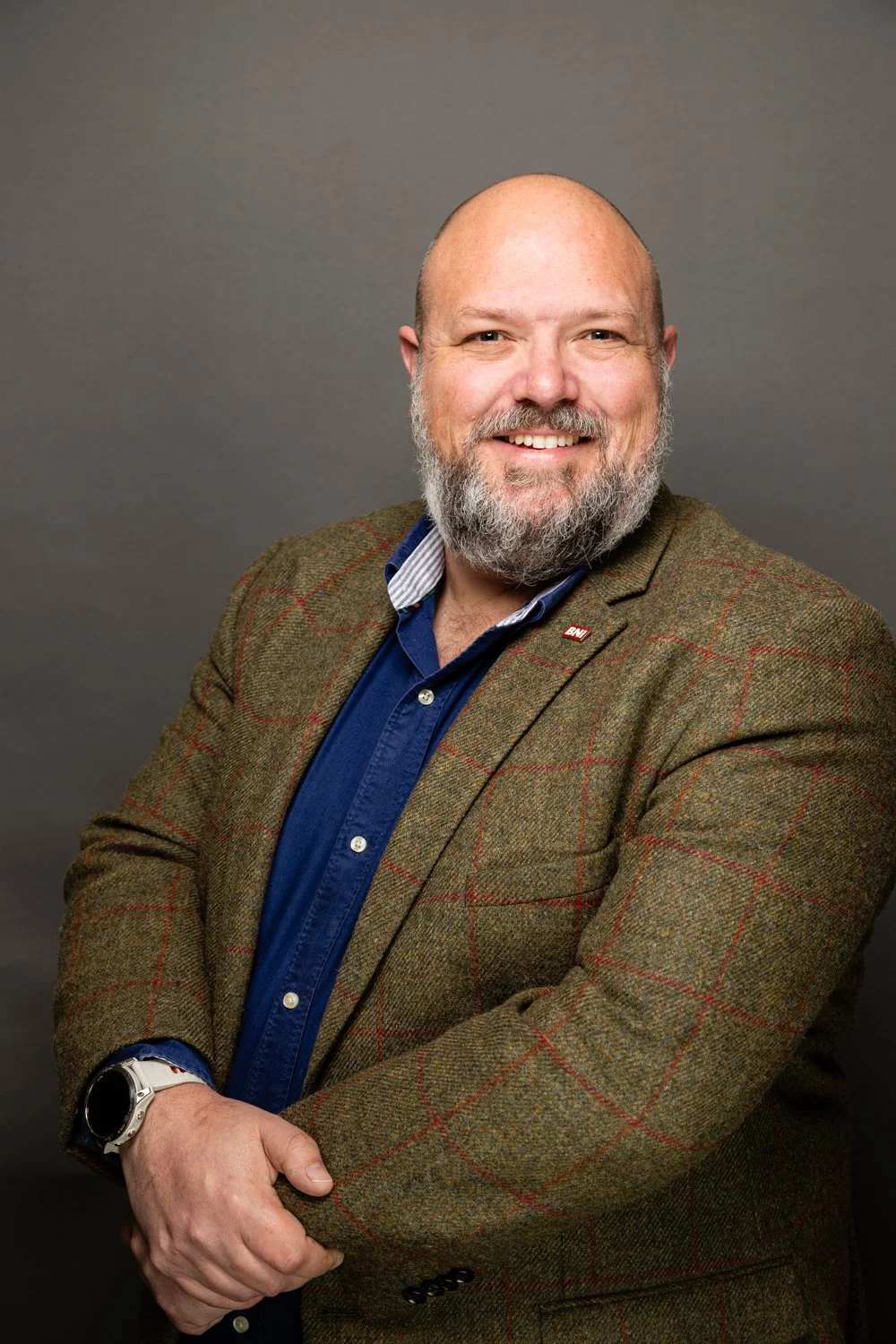 A smiling man with a beard and bald head wearing a brown checkered blazer over a blue shirt, posing against a plain gray background.