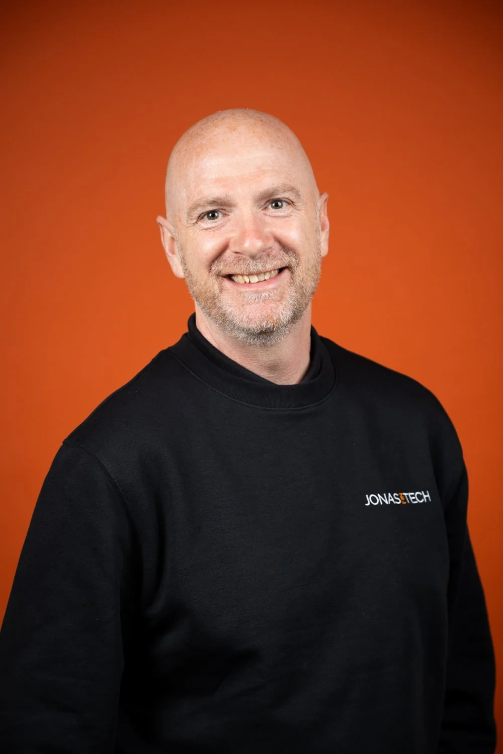 A smiling man with a bald head and beard wearing a black Jonastech sweatshirt, standing against an orange background.