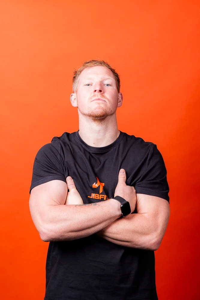 A man with light skin and short reddish hair standing against an orange background. He is wearing a black T-shirt with a logo on it, a watch on his left wrist, and has his arms crossed, with a serious expression.