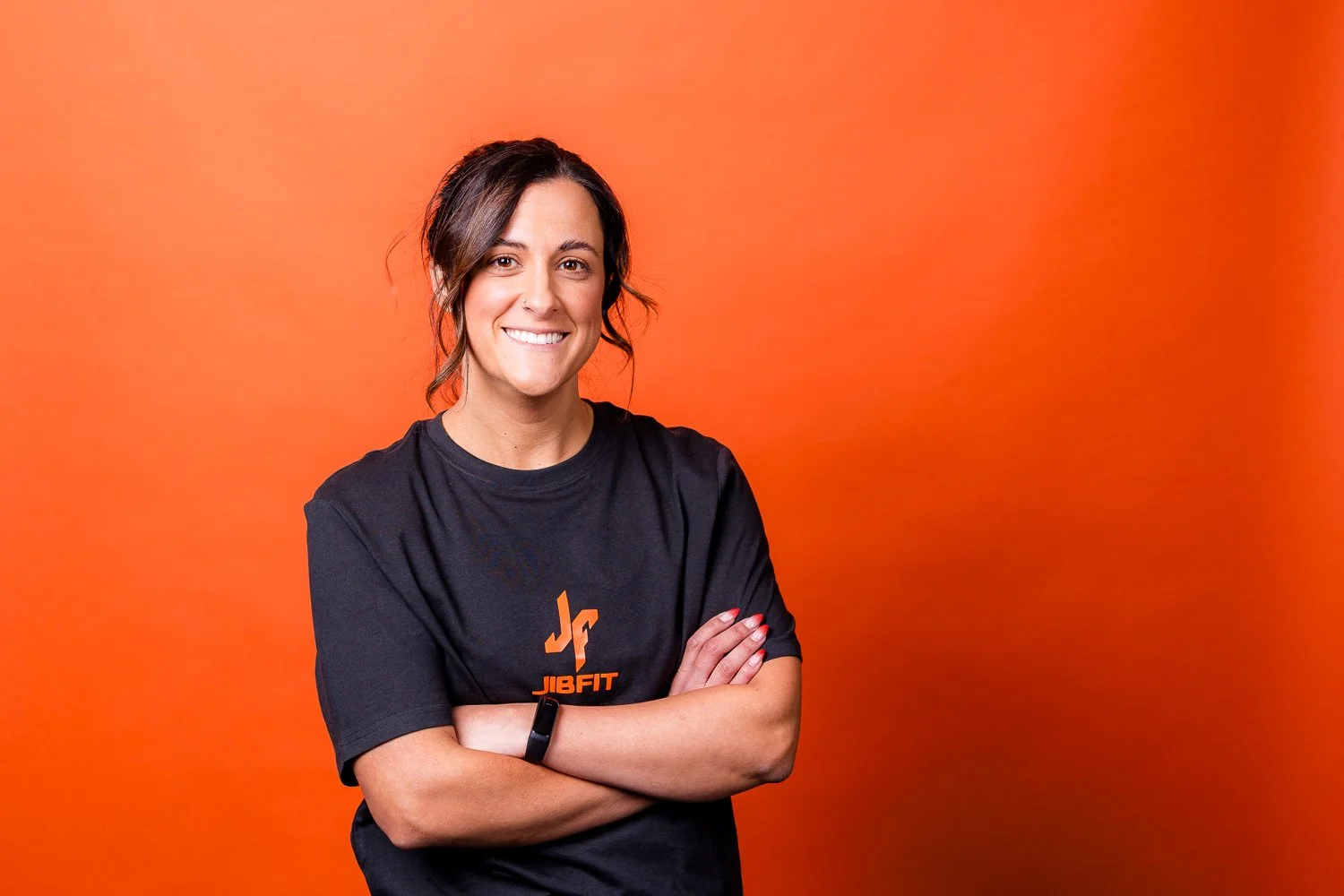 Smiling woman with dark hair tied back, crossing her arms, wearing a black T-shirt with orange logo and text, standing against an orange gradient background.
