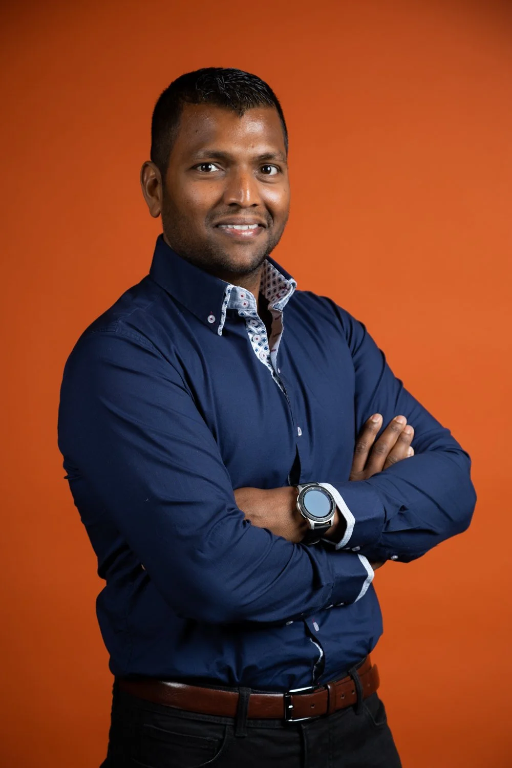 A man with dark hair and brown skin, wearing a dark blue button-up shirt with a patterned interior collar, standing with arms crossed, against an orange background.