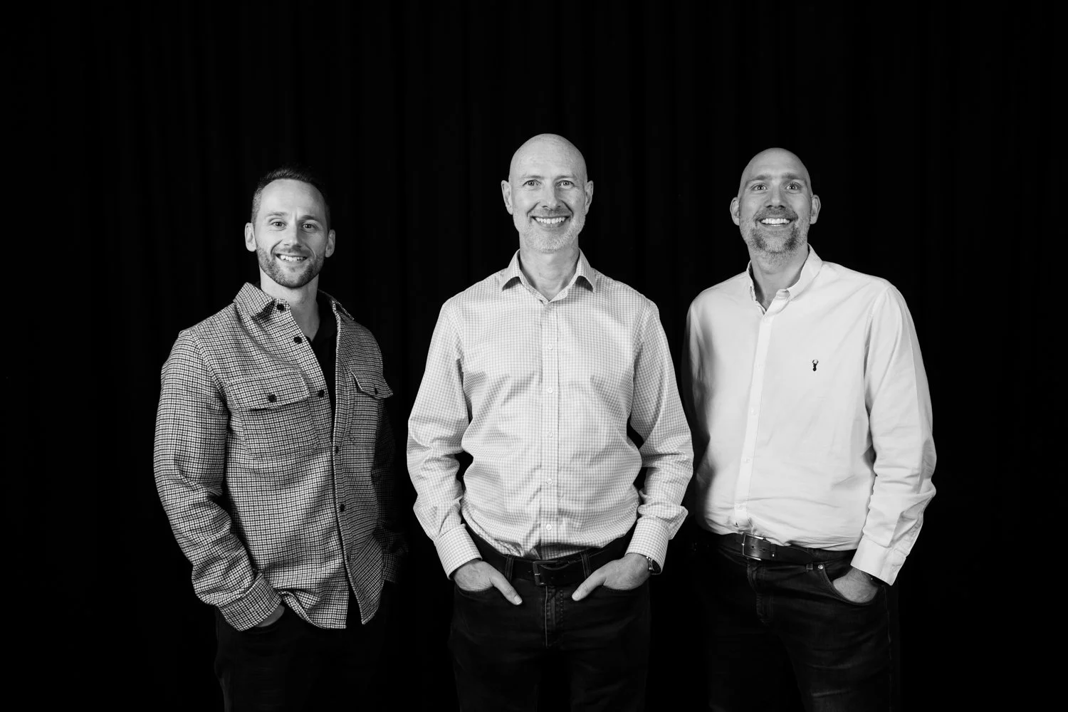 Black and white photo of three men standing with hands in pockets against a dark background, smiling at the camera.