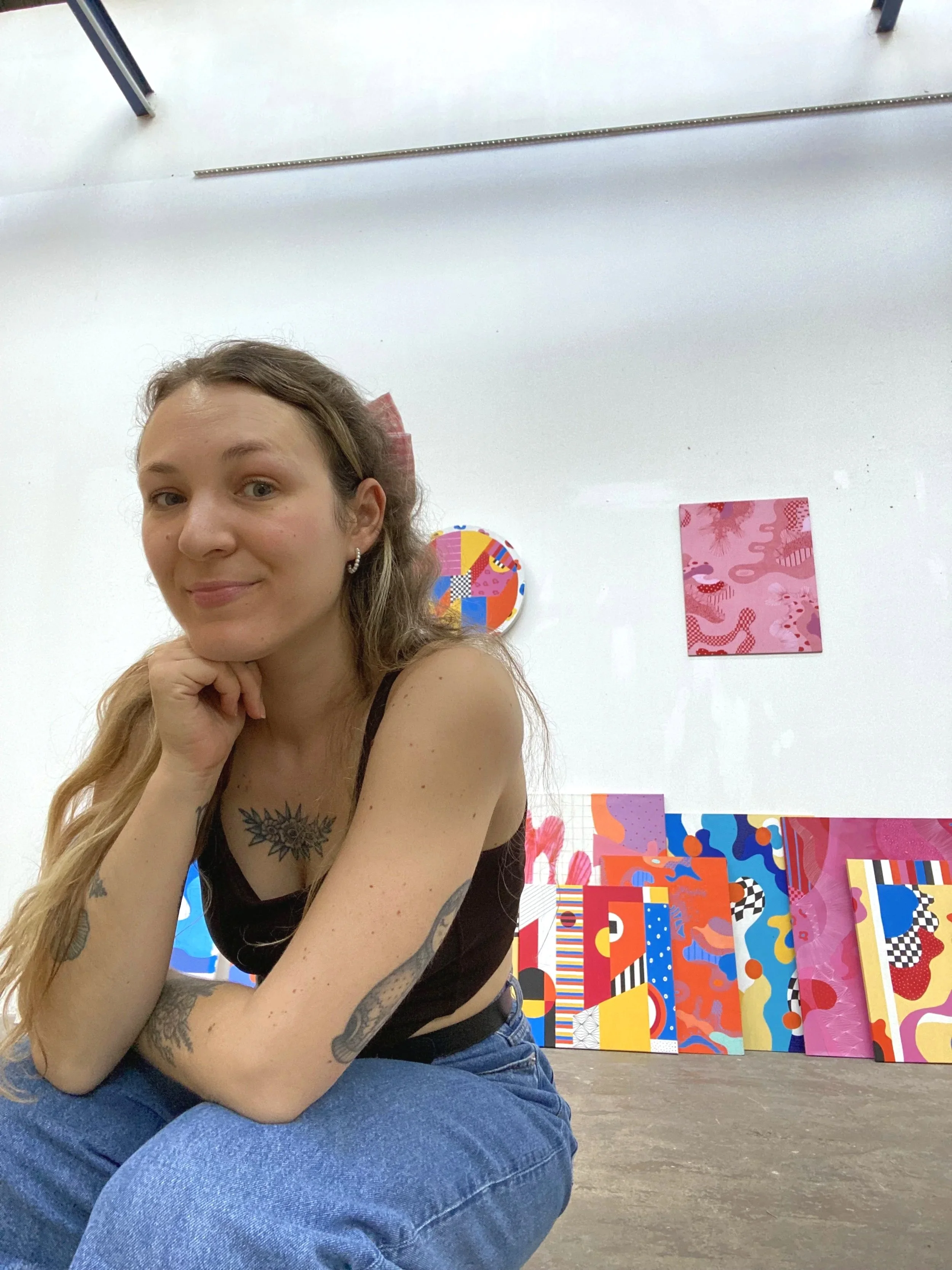 Jeune femme avec des tatouages, assise au sol, souriant, autour d'œuvres d'art modernes et colorées.