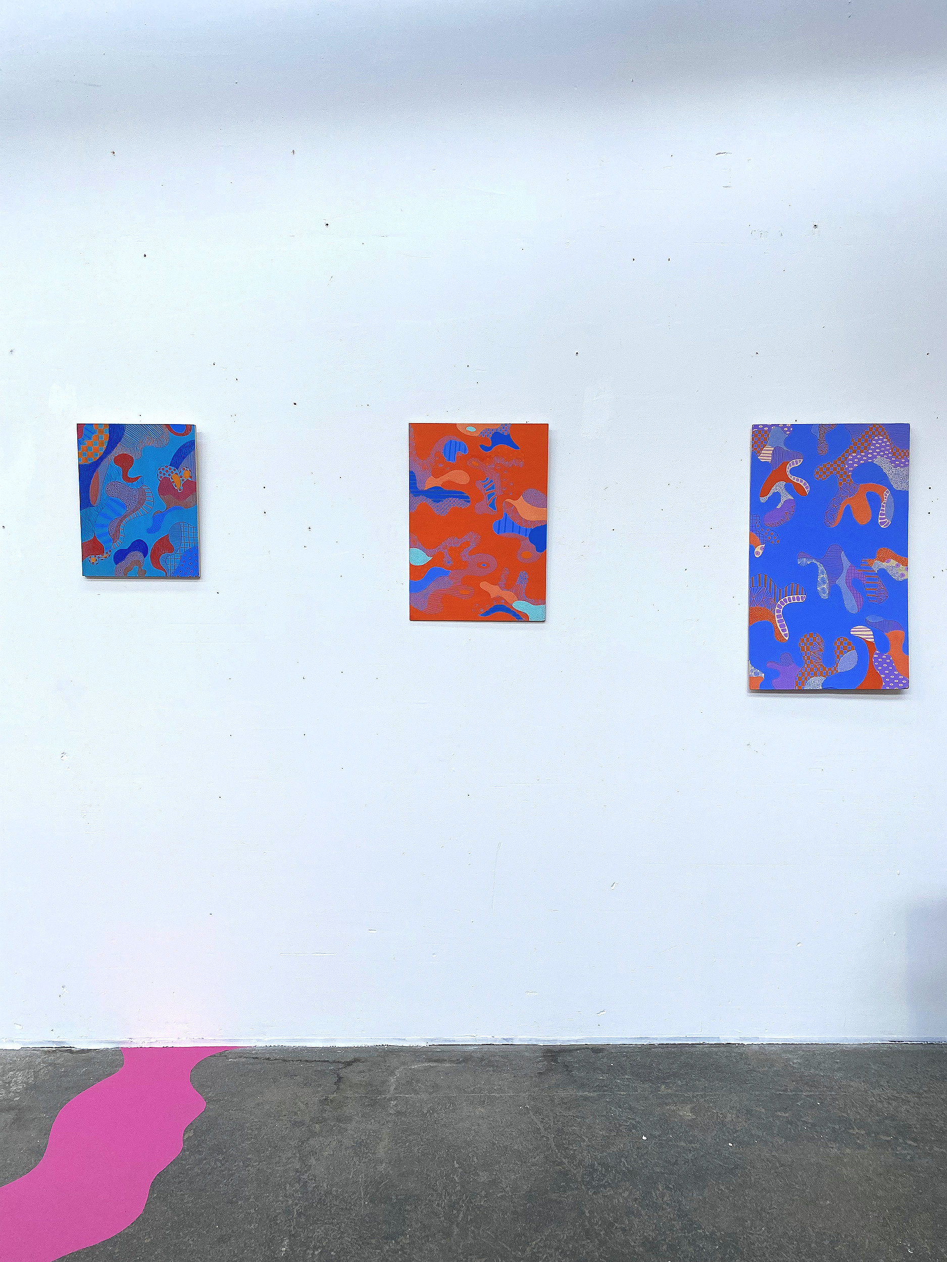 Trois tableaux abstraits avec motifs colorés orange, bleu, violet et rouge accrochés sur un mur blanc dans une galerie d'art.