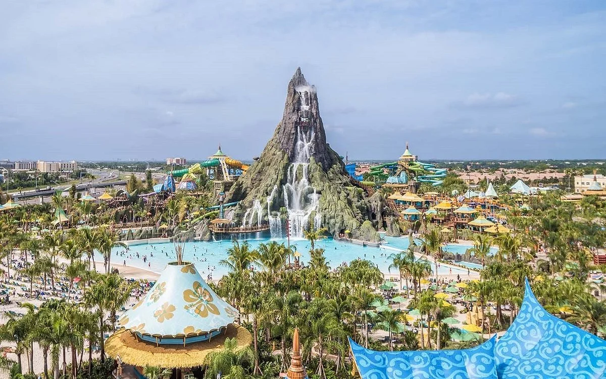 Large water park with a volcano-themed water slide structure, pools, and numerous water slides and attractions in the background, surrounded by palm trees.