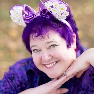 A smiling woman with her hands under her chin with purple hair and purple Mickey ears