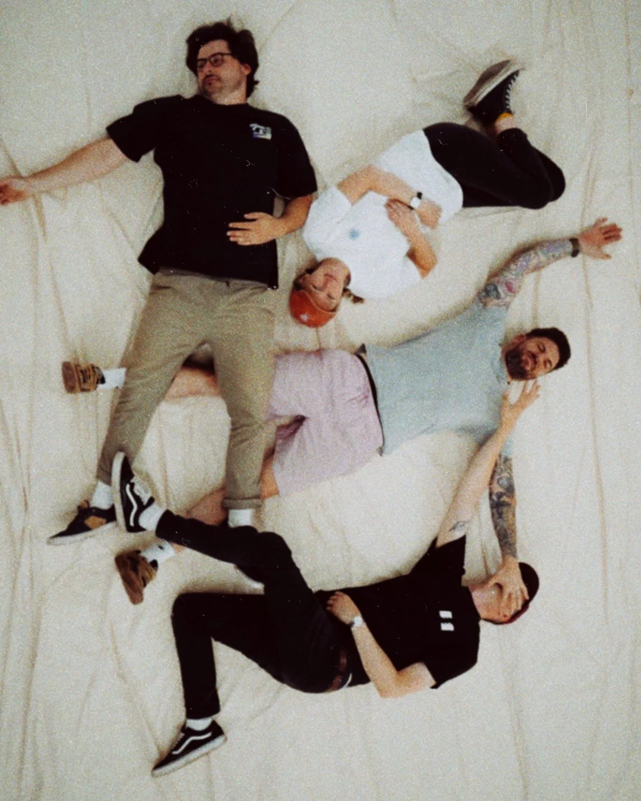 Four people lying on a beige floor forming a star shape, with their heads toward the center and arms and legs extended outward.