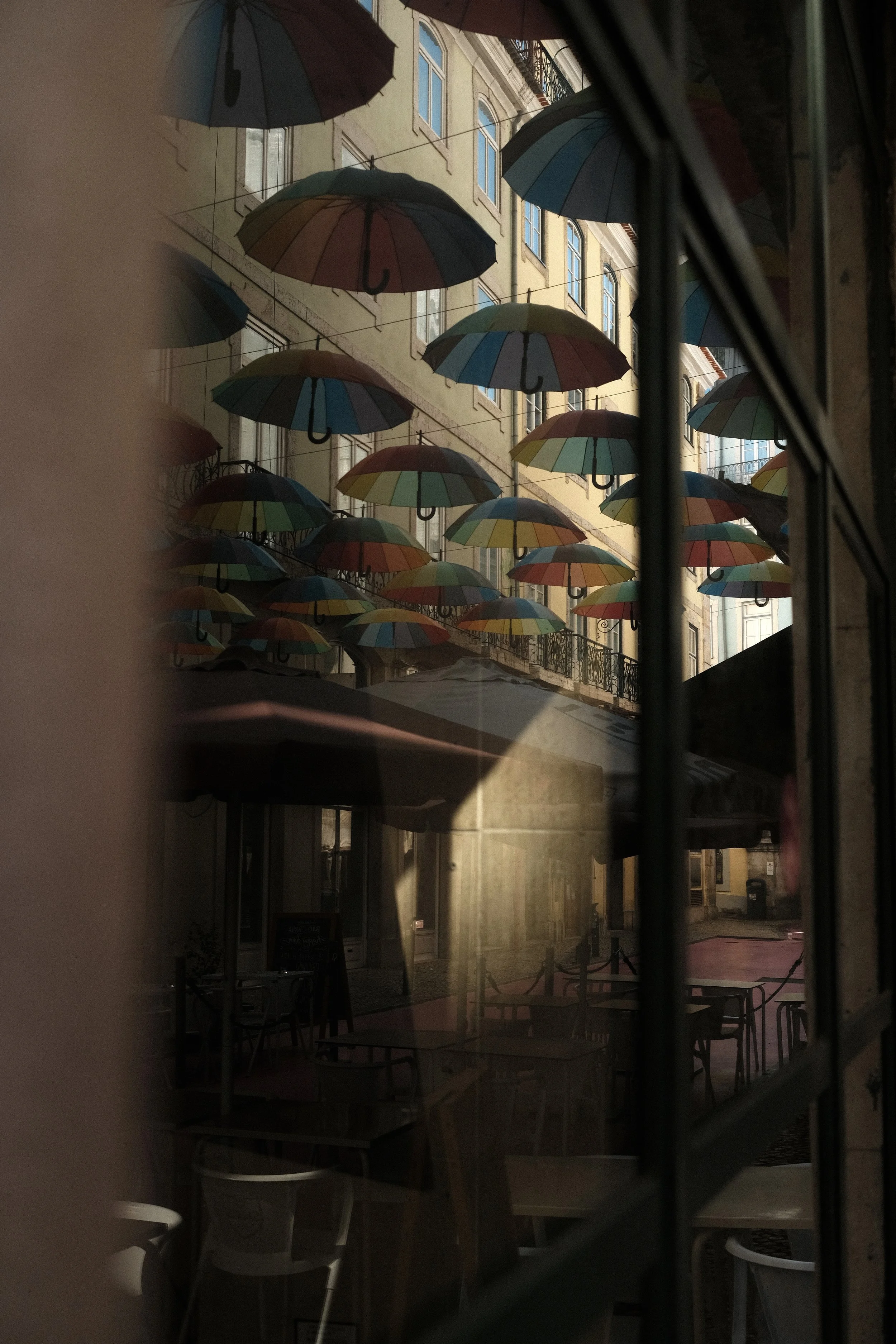 Viewing through a window, a street scene with numerous colorful umbrellas hanging overhead, creating a canopy above outdoor cafe tables.