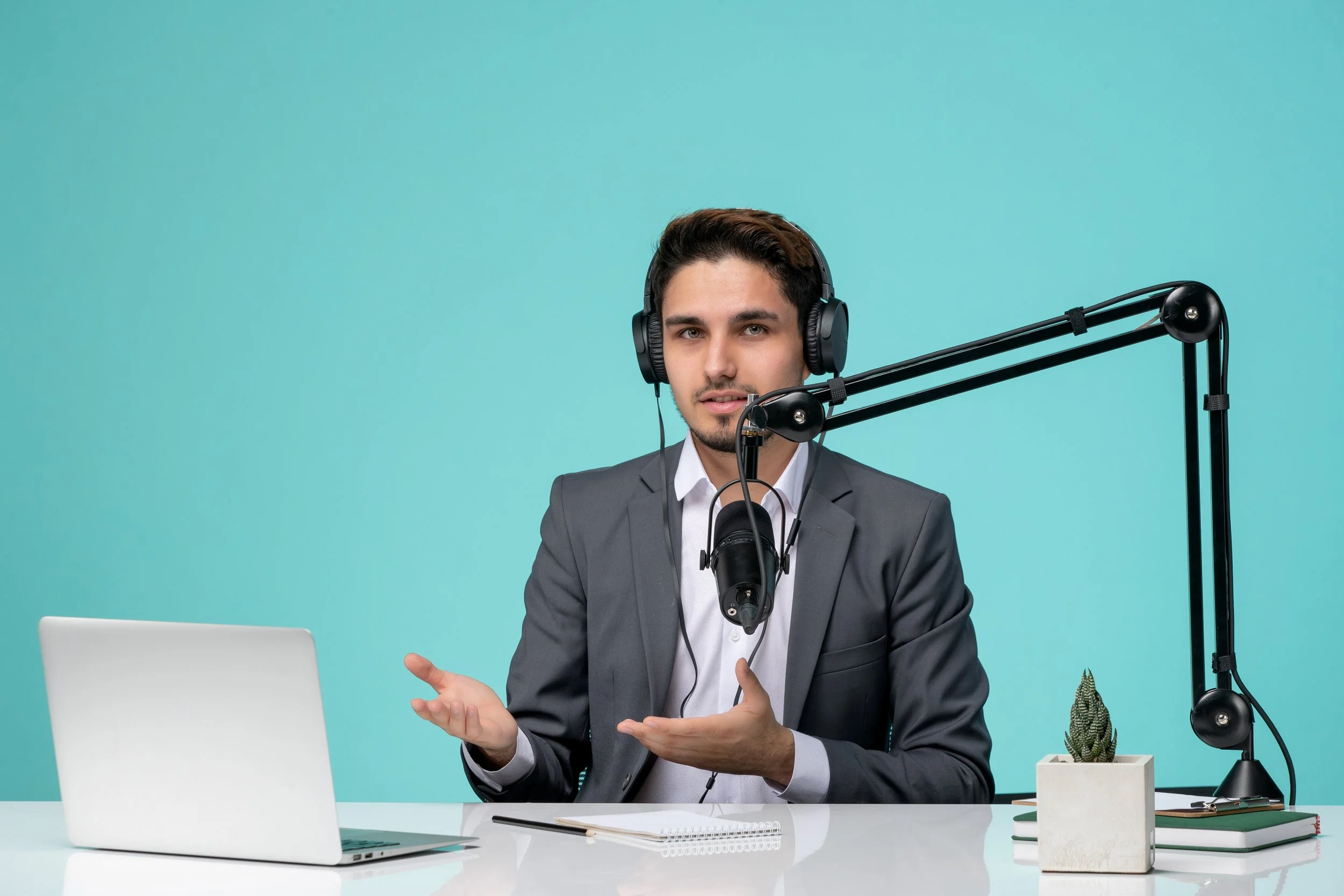 A man in a business suit wearing headphones, speaking into a microphone during a podcast or radio show, seated at a white desk with a laptop, notebook, small potted plant, and books against a turquoise background.