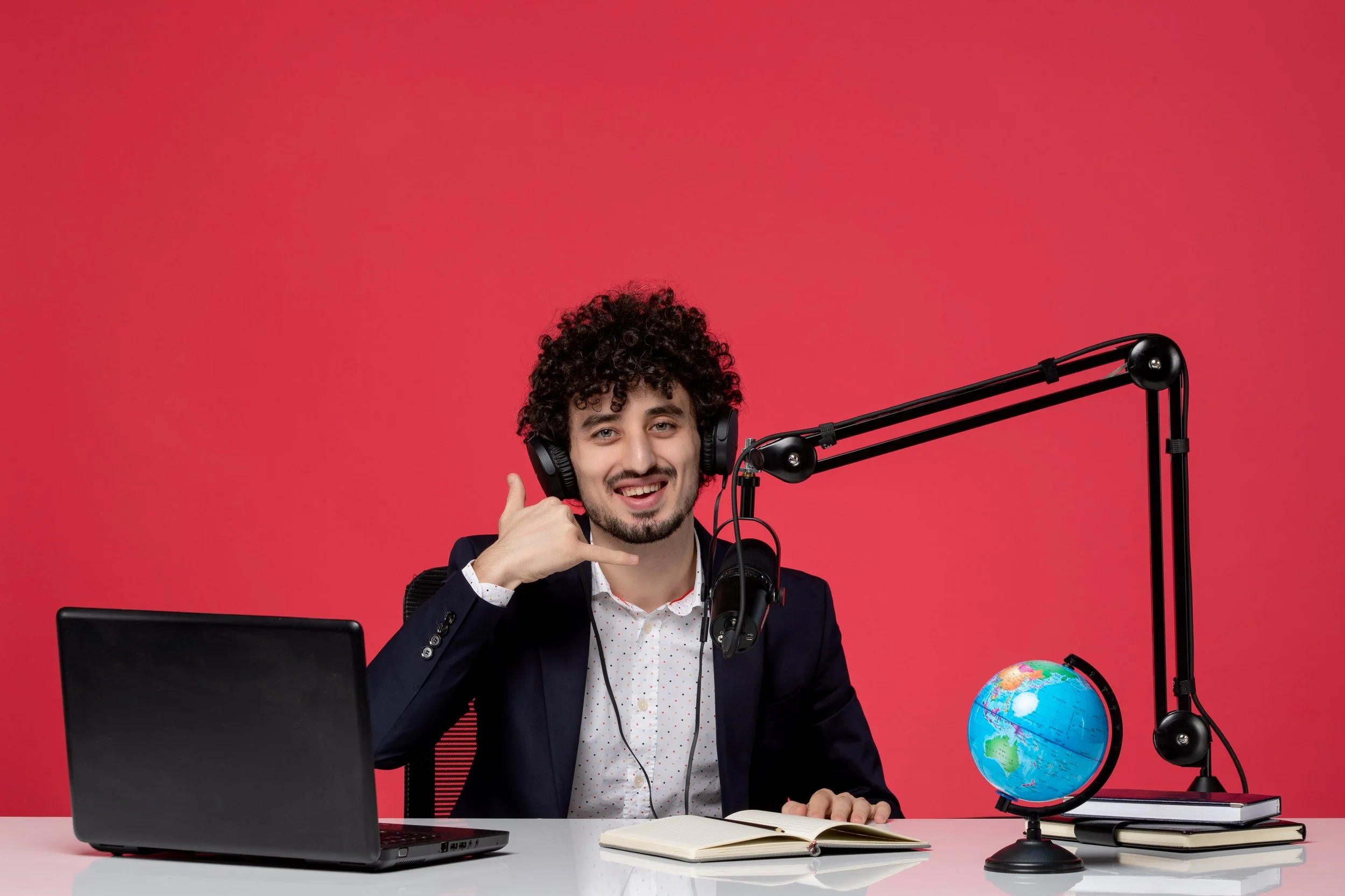 A man with curly hair wearing headphones, seated at a desk with a laptop, books, a globe, and a microphone, making a phone gesture with his hand, smiling, against a red background.