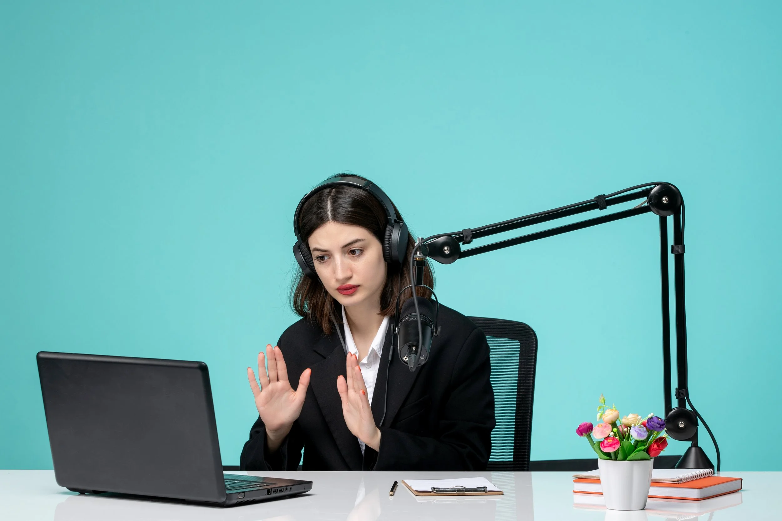 A woman with dark hair, wearing a black blazer and white shirt, sits at a desk with a laptop, headphones, and a large adjustable microphone arm. She appears to be engaged in a video call or recording. There is a small bouquet of colorful flowers and notebooks on the desk against a plain turquoise background.