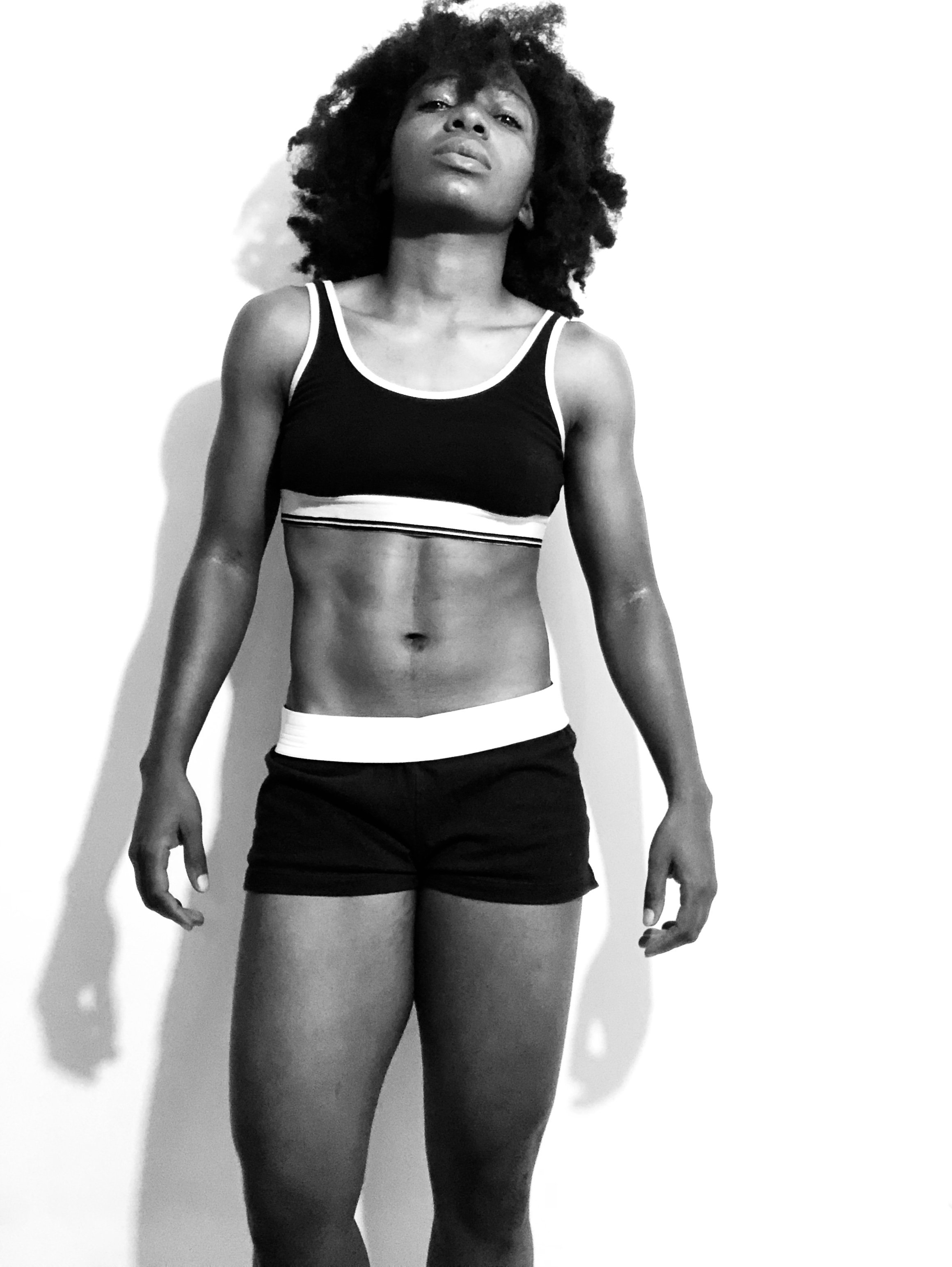 A black and white photo of a young woman with curly hair in athletic wear, standing against a white wall, looking confidently at the camera.