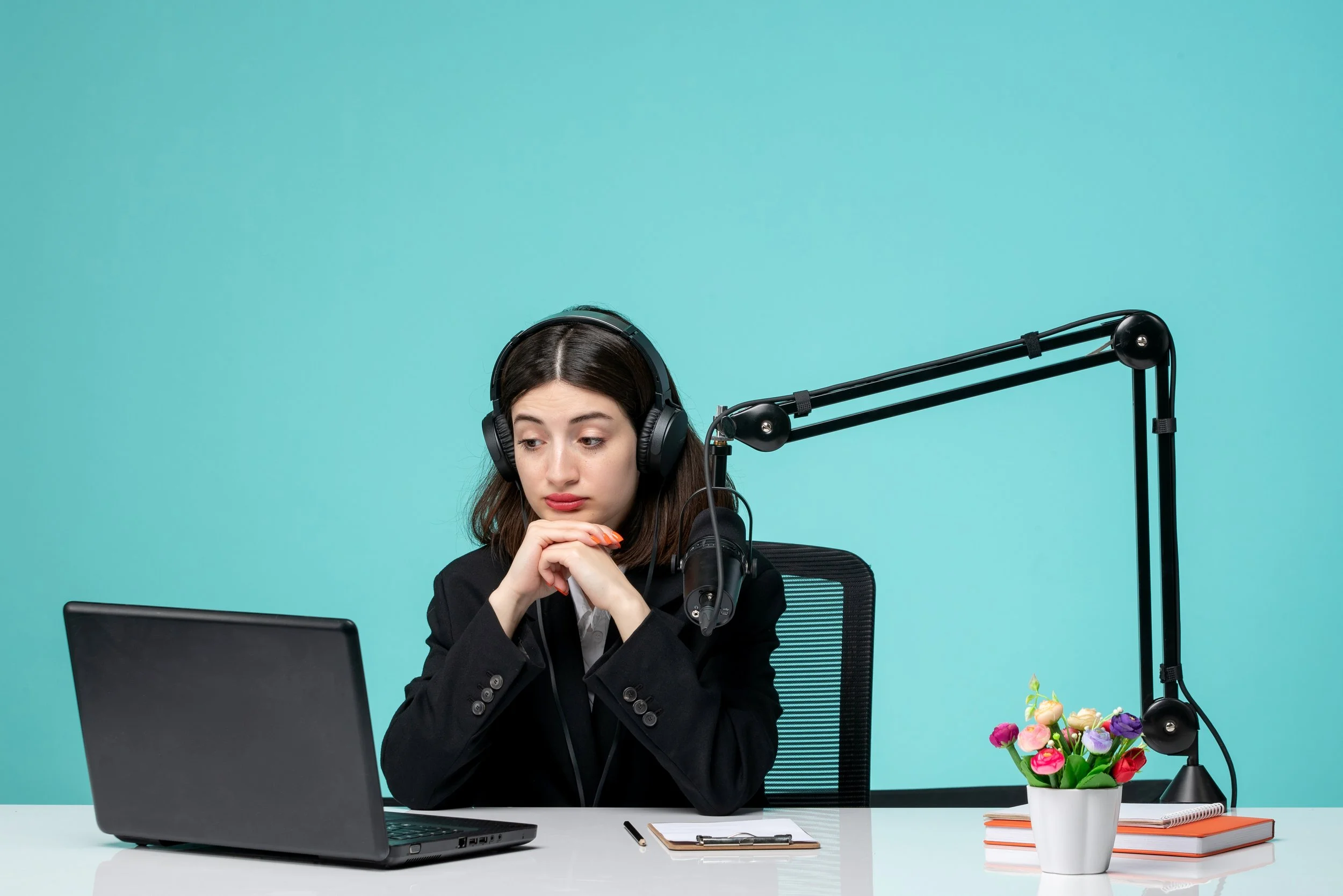 A woman in business attire sitting at a desk with a laptop, wearing large headphones, looking thoughtful, with a microphone connected to an adjustable arm, and a small potted plant with colorful flowers.