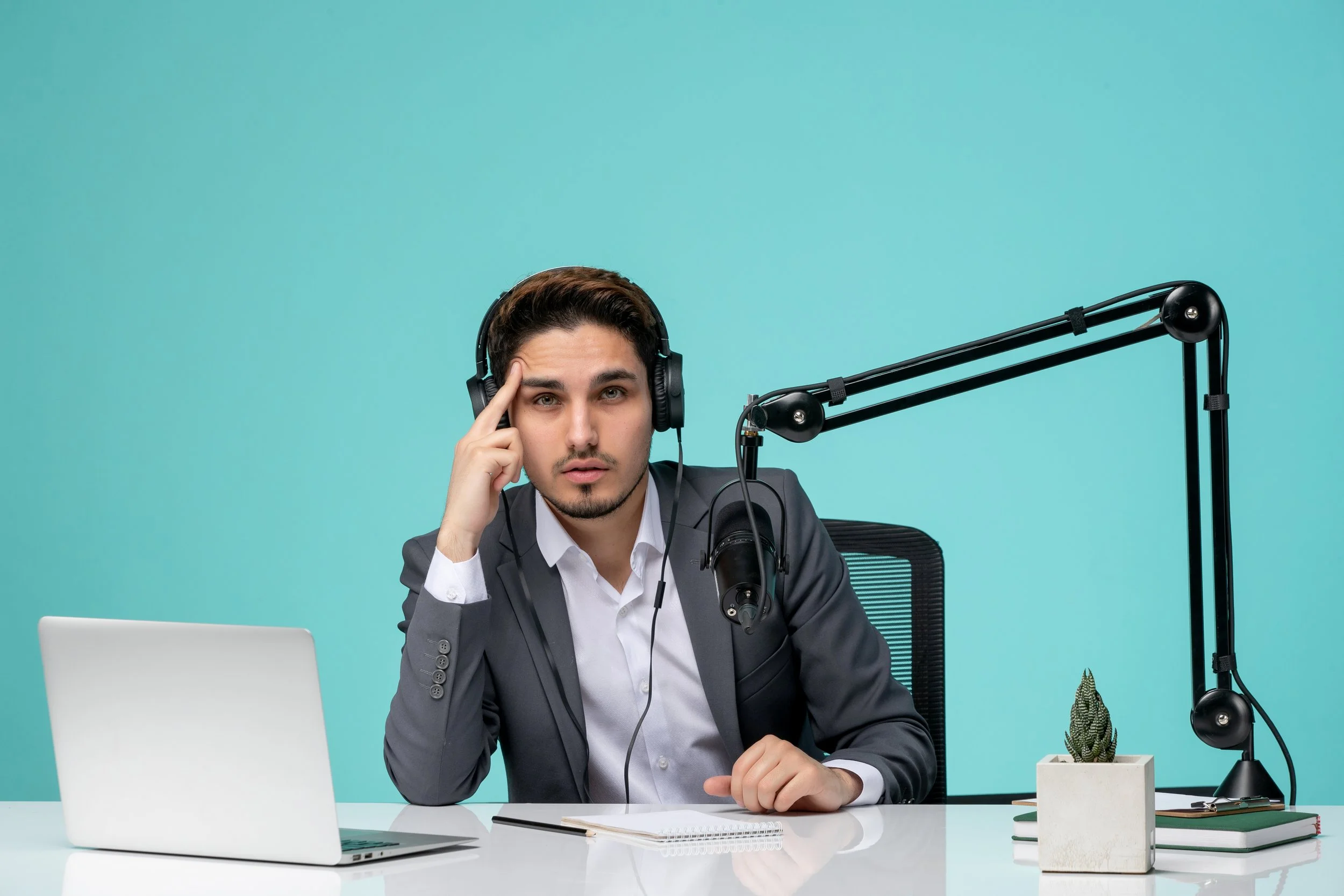A man in a gray suit and white shirt sitting at a desk with a laptop, notebook, and a small potted plant, wearing headphones, with a serious expression, against a plain teal background.