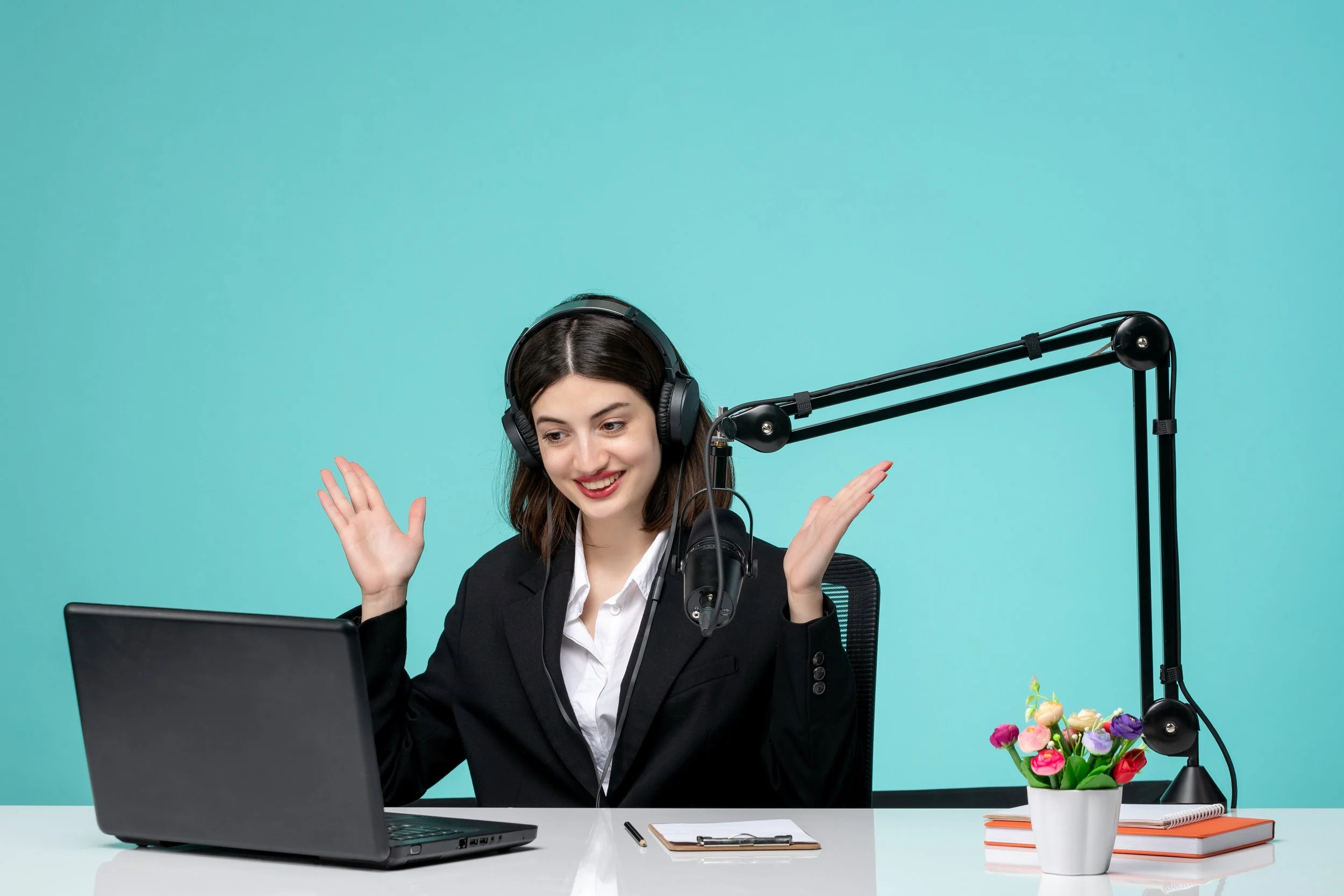 A woman in a black blazer and white shirt sitting at a desk, wearing large headphones, smiling, with her hands raised in celebration, in front of a laptop, with a notebook, pen, and potted flowers on the desk, against a teal background.