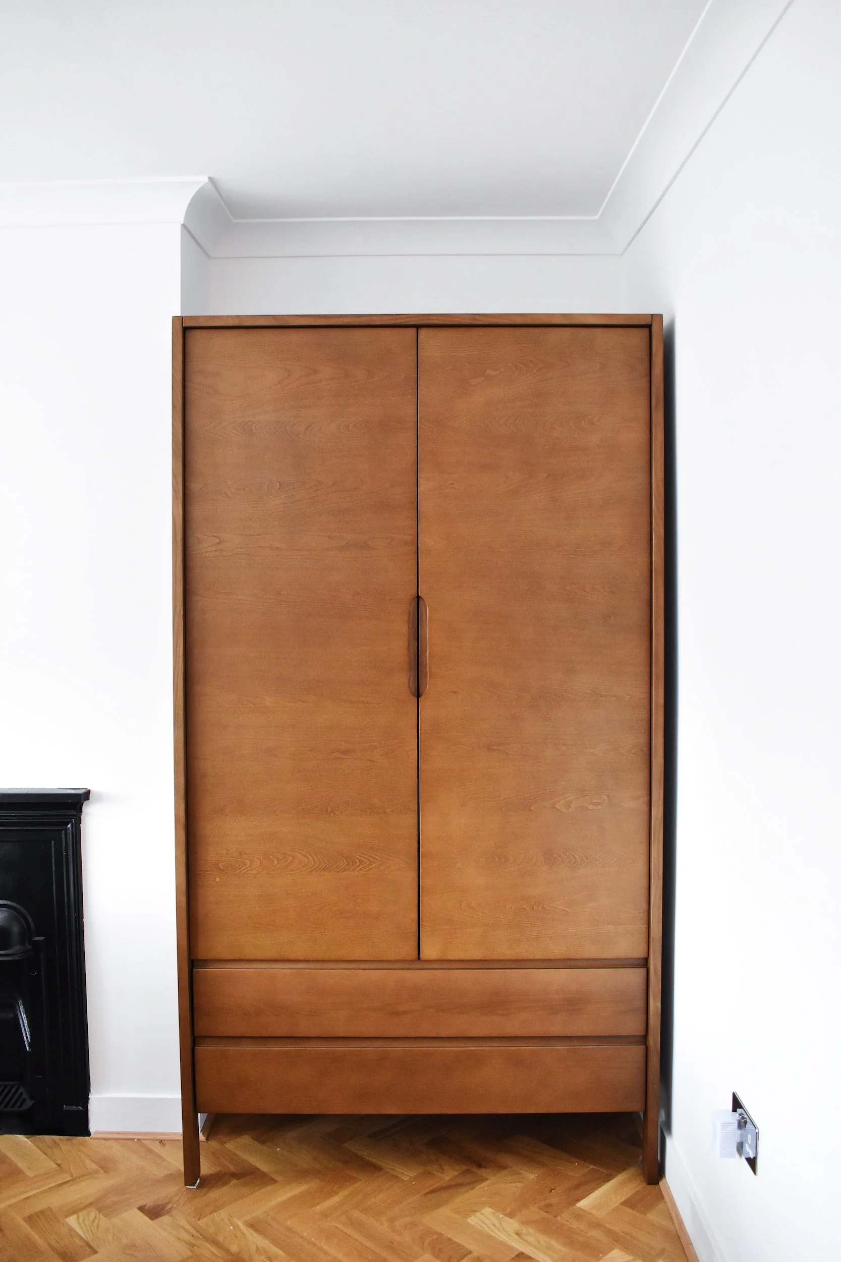 A wooden wardrobe with two doors and two drawers, standing against a white wall in a room with parquet flooring.