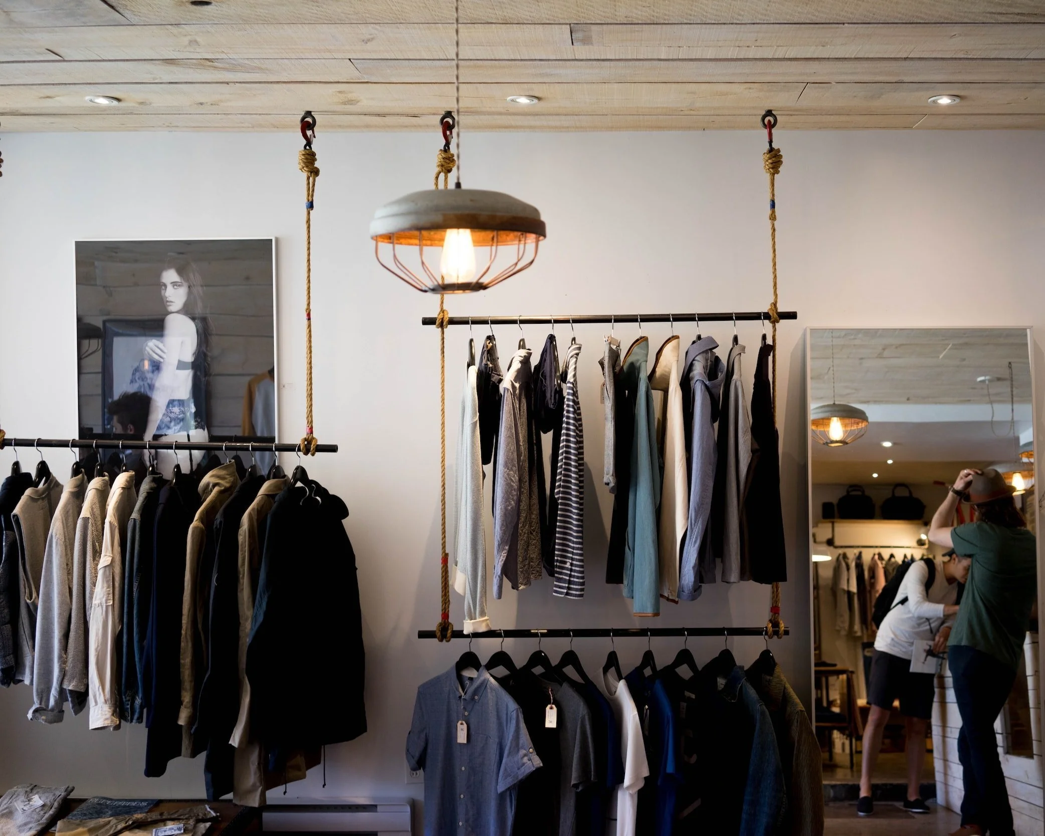 Clothing store interior with racks of clothes, a hanging light fixture, a mirror reflecting people, and a black-and-white photograph on the wall.