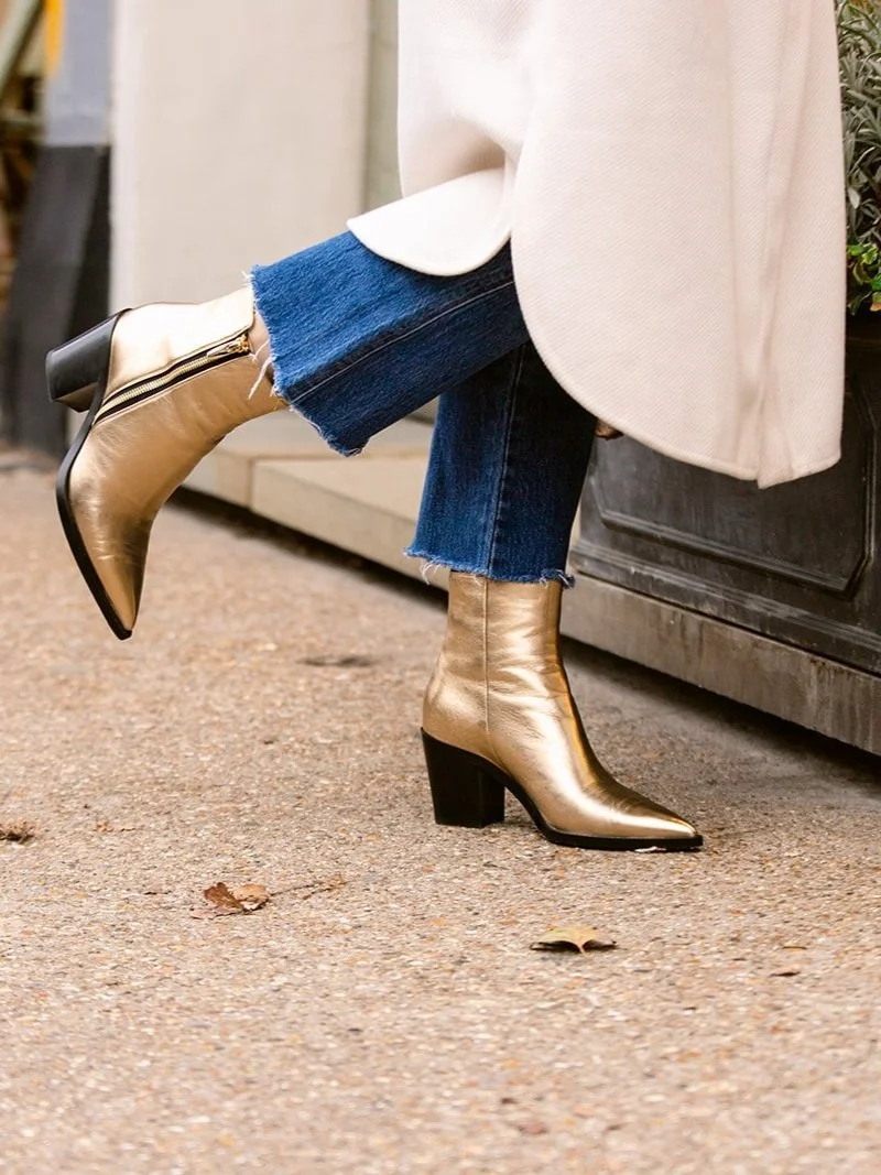 Person wearing gold ankle boots, blue jeans, and a white coat, standing on a sidewalk with fall leaves.