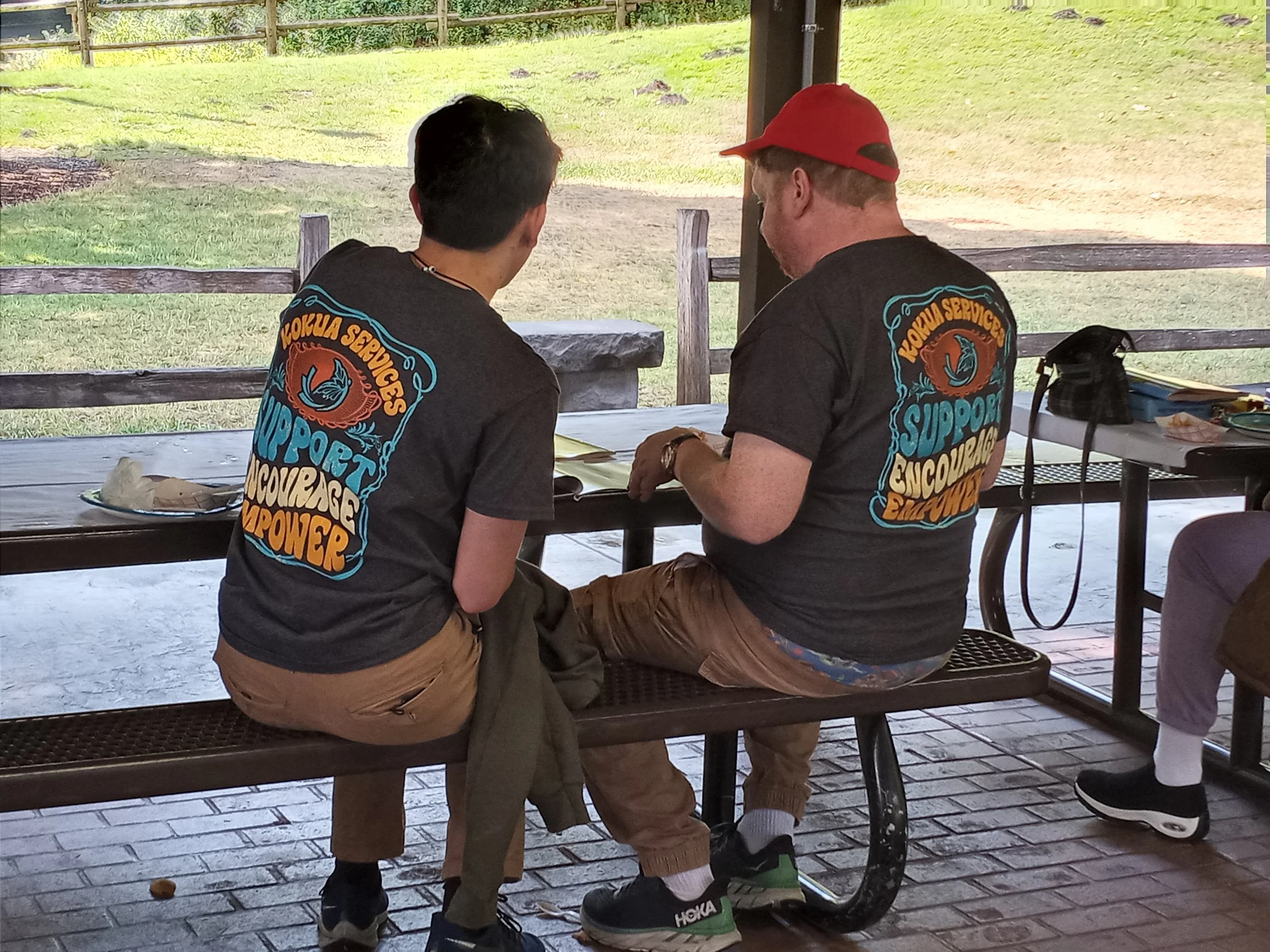 Two young men sitting on a park bench, wearing matching gray t-shirts with colorful text on the back, talking to each other at a picnic shelter. One is wearing tan pants, the other is in camouflage shorts with a red cap, and they have various items on the table behind them.