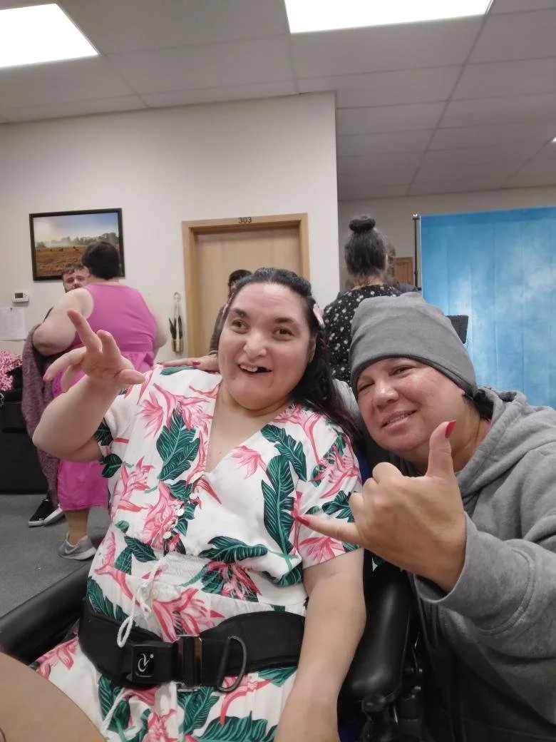 Two people sitting together, smiling and making hand gestures, in a room with other people and a blue backdrop.