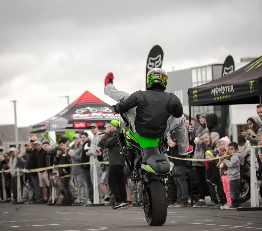 A motorcyclist performing a wheelie on a green and black motorcycle at a crowded outdoor event with spectators and tents.