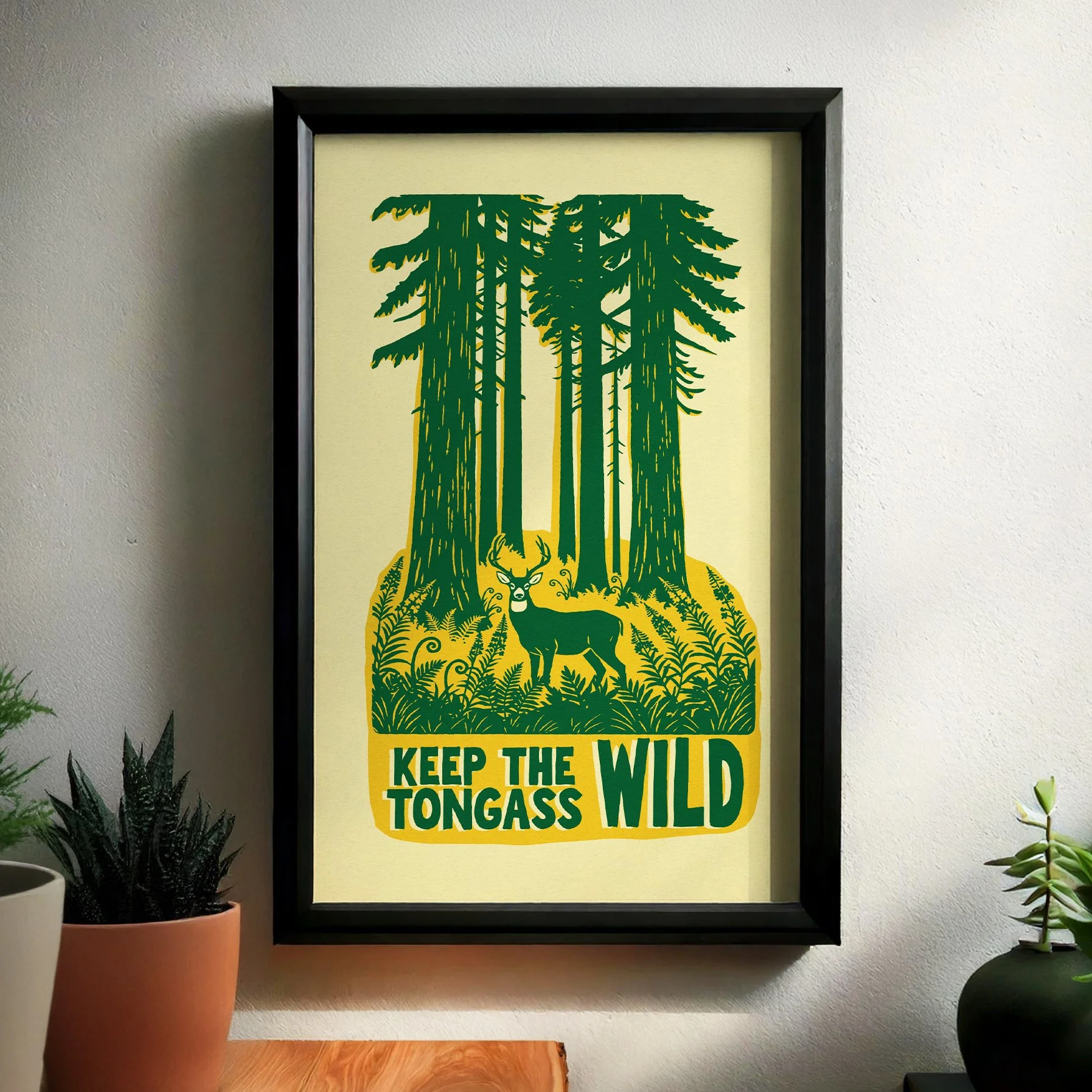 Framed ‘Keep the Tongass Wild’ screen print hanging on a light-colored wall. The artwork shows tall green spruce trees, lush ferns, and a black-tailed deer in a two-color vintage-style design.