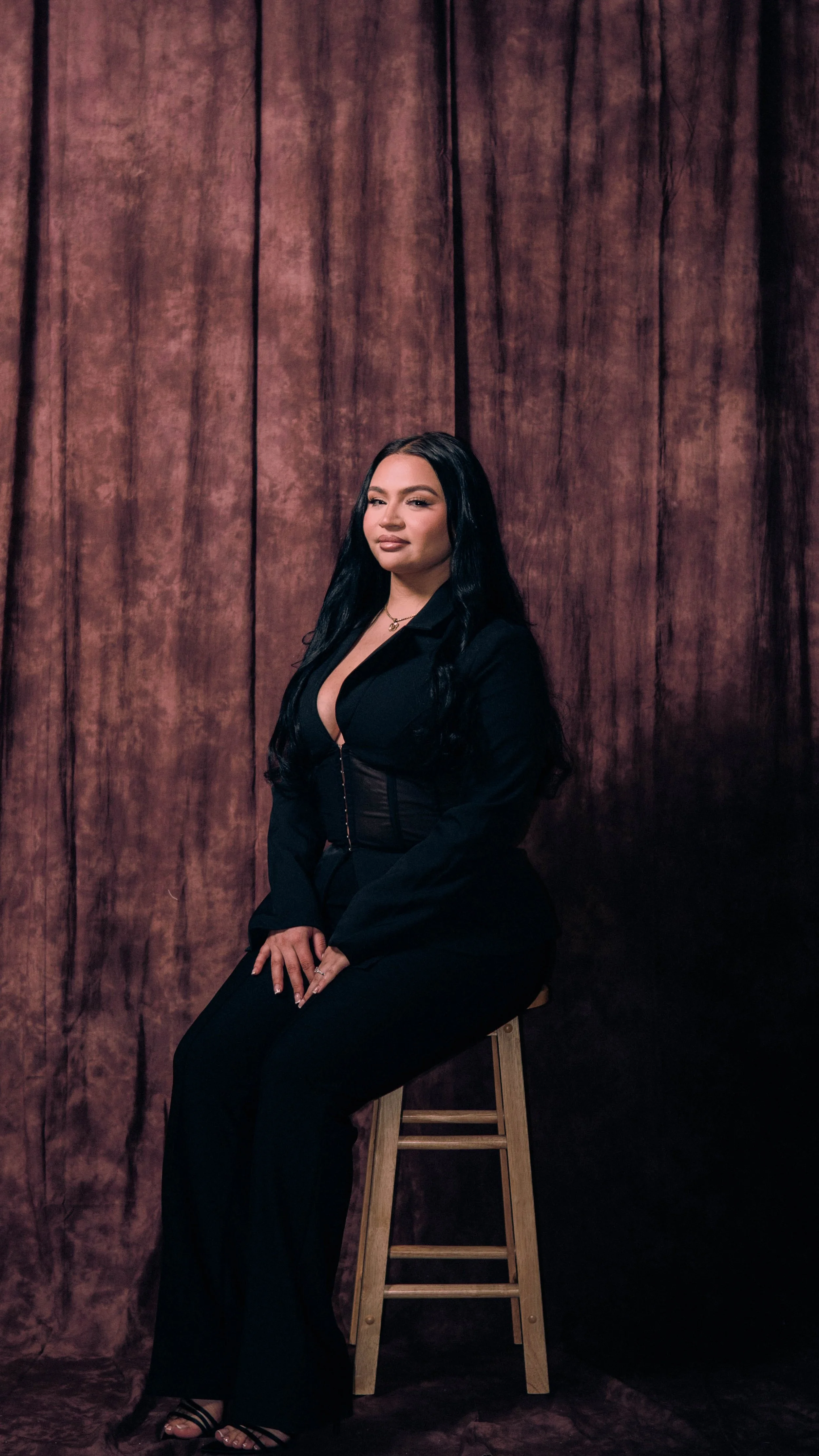 A woman with long black hair sitting on a wooden stool in front of a brown textured curtain, wearing a black blazer and pants, with makeup and jewelry.