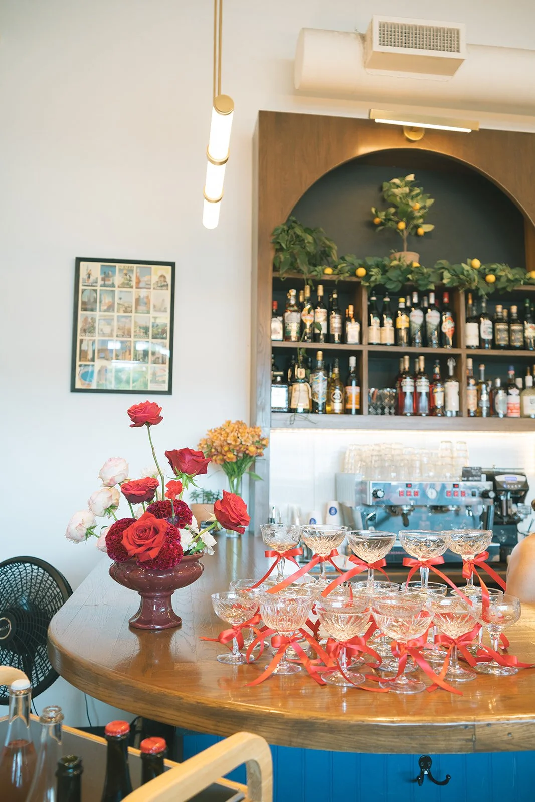 Small goblets with bright and delicate red bows tied on their stems sit stacked on wooden bar, a vase of tall blooming red roses beside it.