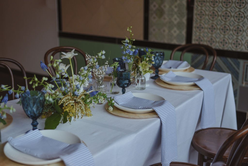 End of a table with a white tablecloth, sitting on top are blue goblets, plates with striped napkins, and several bouquets of blue, green, and white flowers.