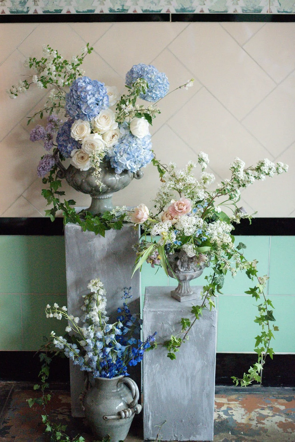 3 vases filled beautifully with white, blue, and green flowers. 2 vases are on pedestals. 