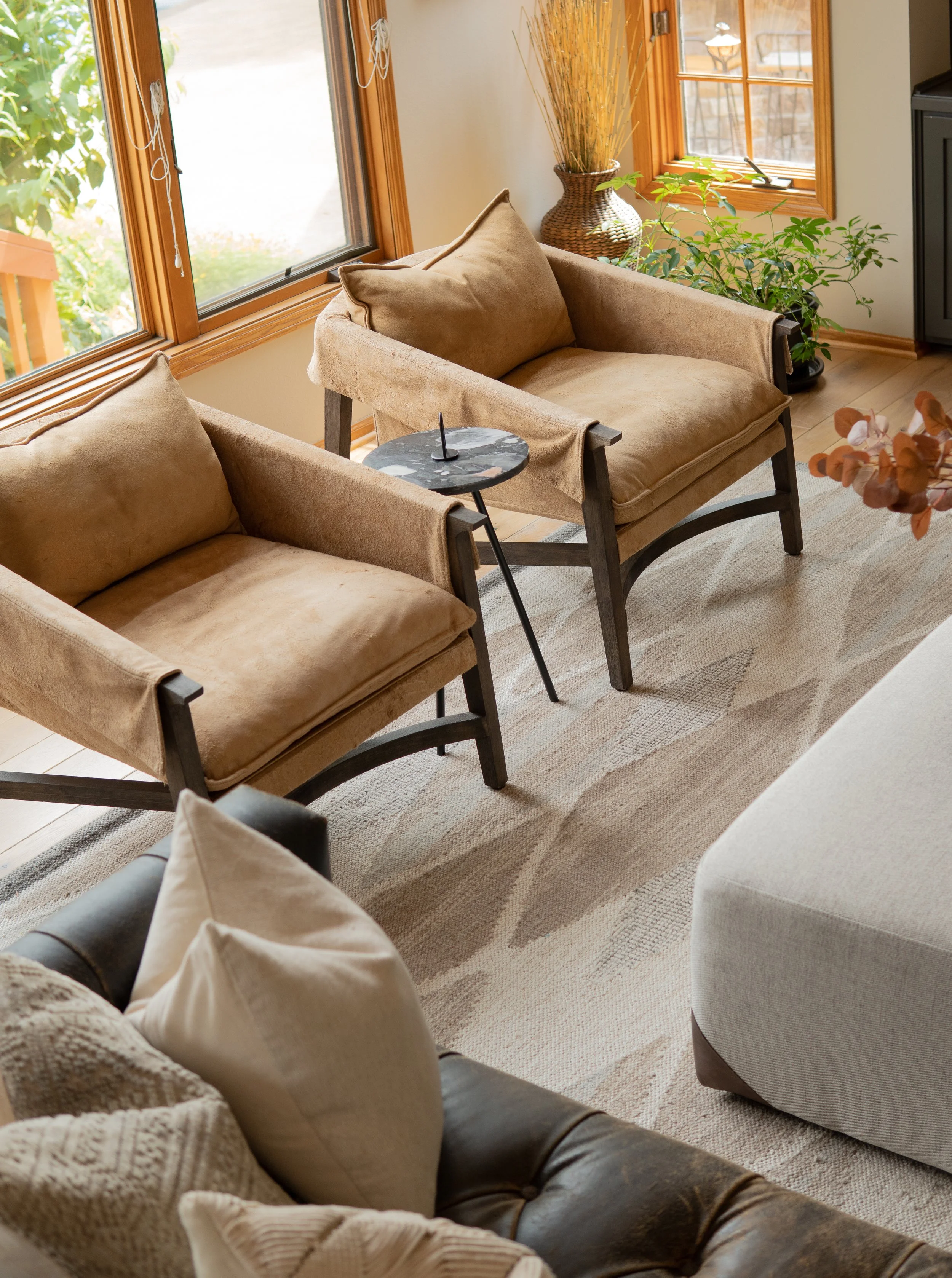 Cozy living room with two beige armchairs, a small round black side table, a light-colored geometric patterned rug, and a woven plant basket near large windows with wooden frames.
