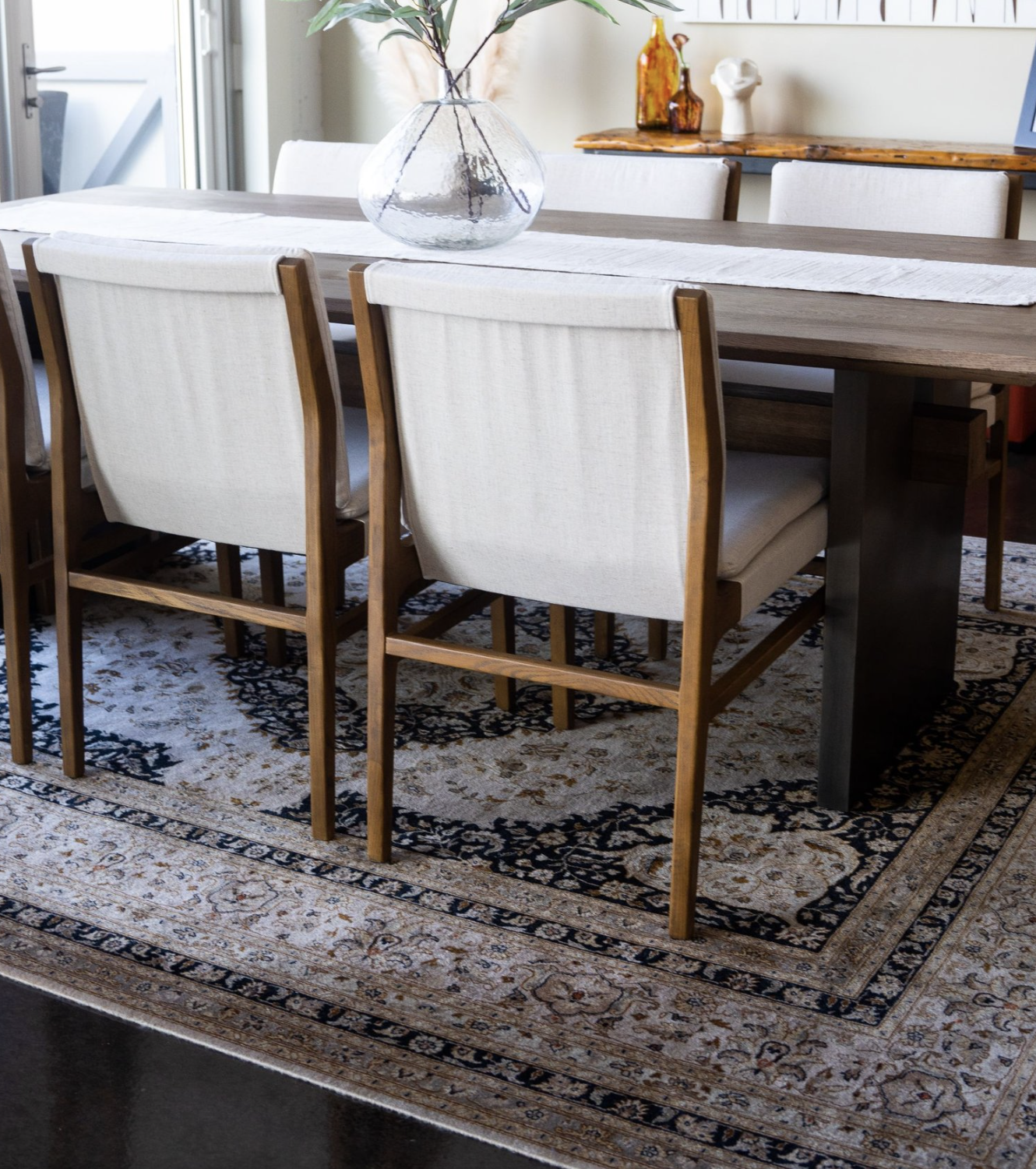 Dining room with wooden table and upholstered chairs, glass vase with green branches, and decorative patterned rug.