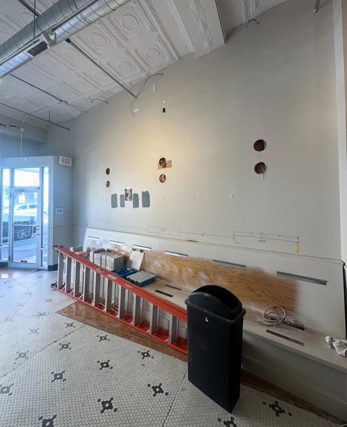 Interior of a building under renovation with a wall that has multiple holes, a red ladder, a black trash can, and construction materials on a bench. The ceiling has decorative molding and exposed ductwork, and the floor features small black and white hexagonal tiles.